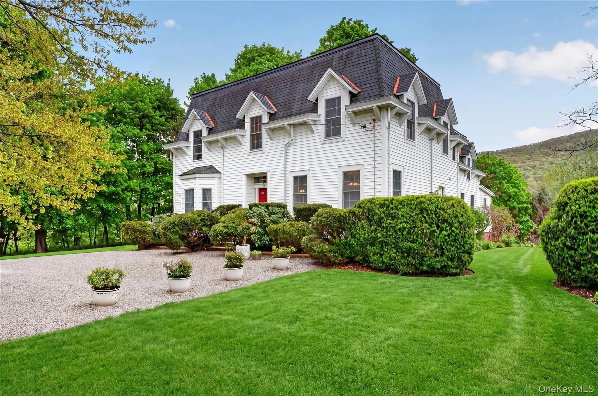 Exterior of colonial residence with red door and mansard roof. Gravel driveway with manicured gardens and lawn surrounding. Exterior of colonial residence with red door and mansard roof. Gravel driveway with manicured gardens and lawn surrounding.