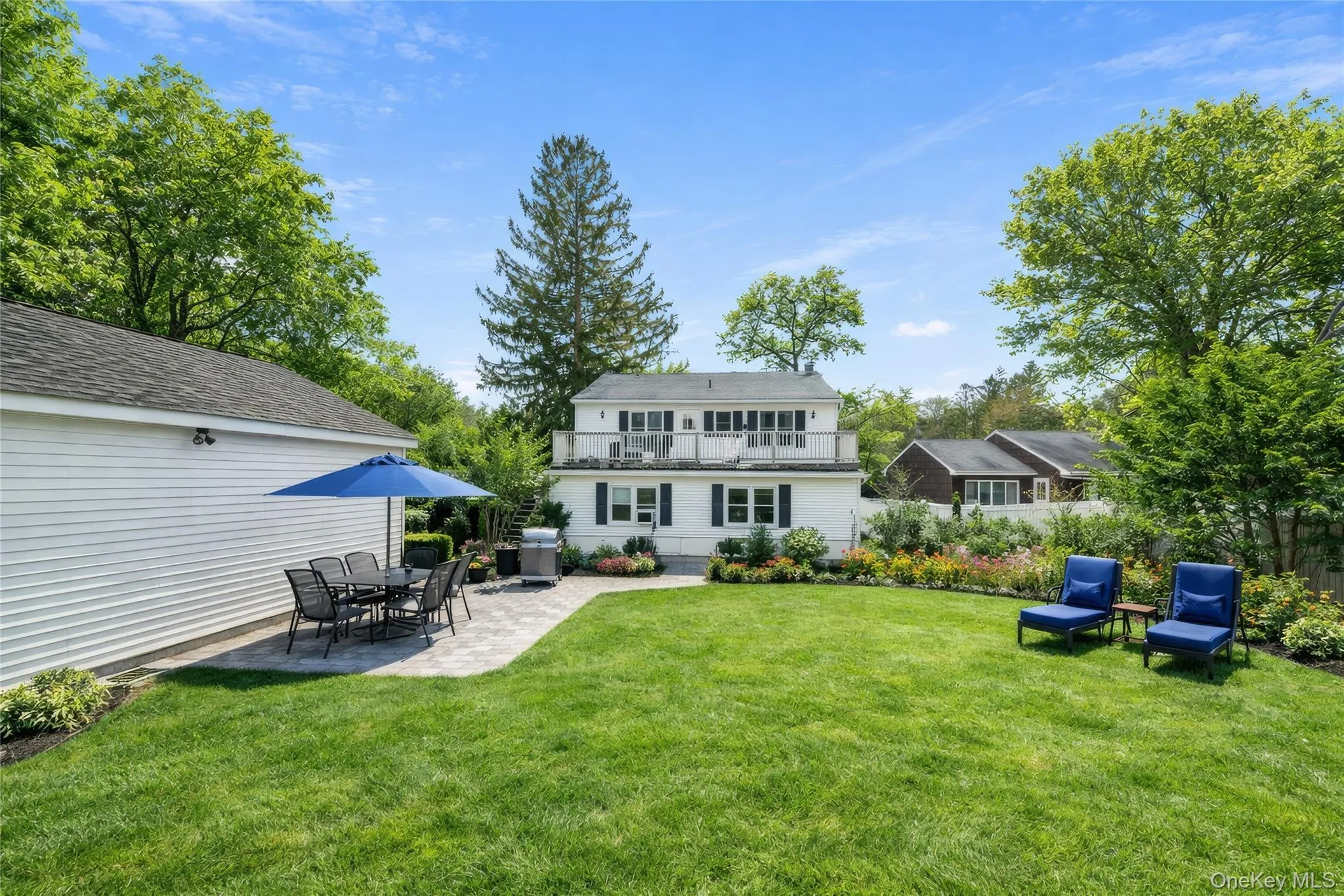 Expansive backyard featuring a large green lawn, a stone paver patio, a white siding garage, and a two-story white residence with dark shutters Expansive backyard featuring a large green lawn, a stone paver patio, a white siding garage, and a two-story white residence with dark shutters
