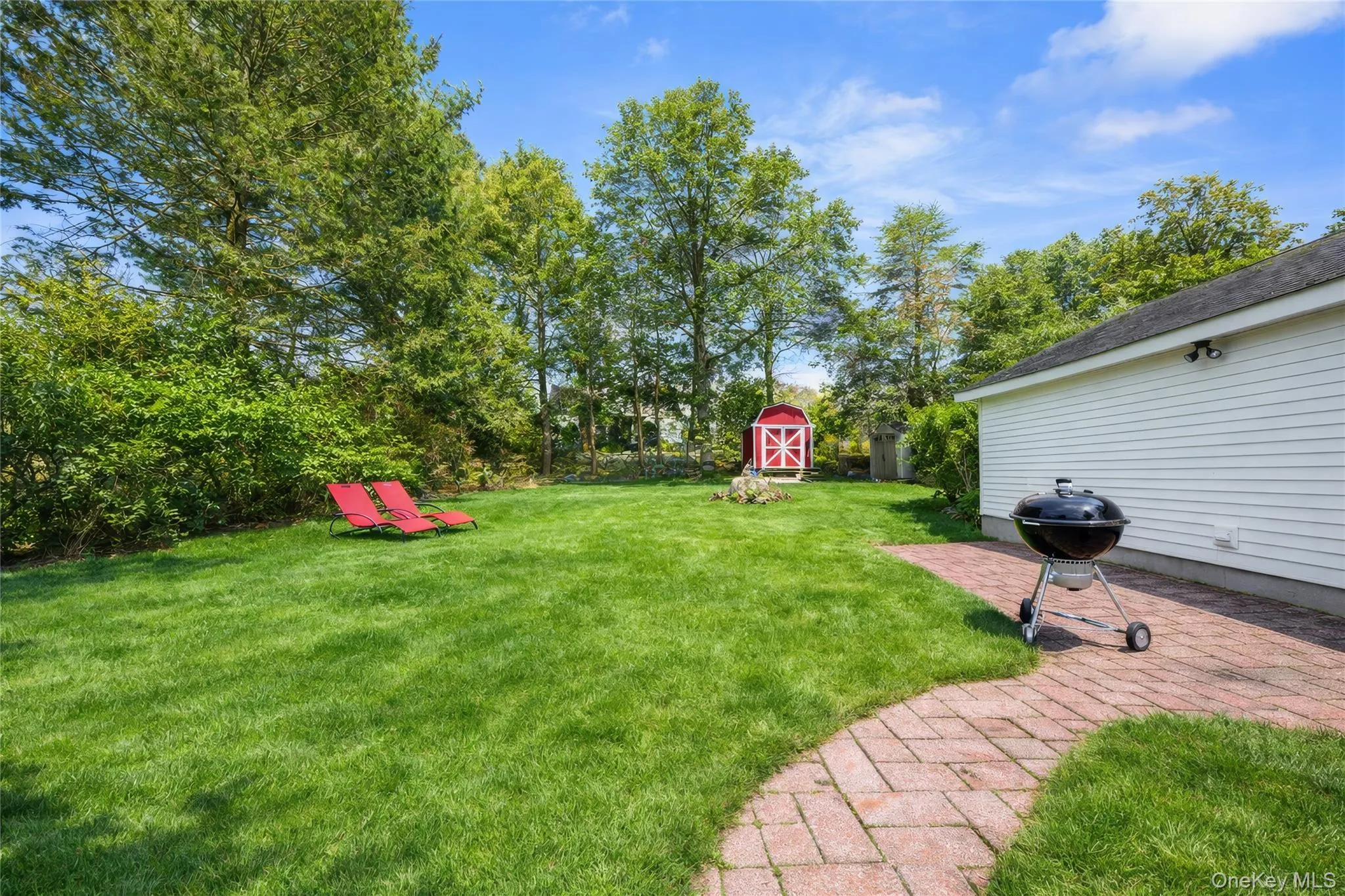 Expansive green lawn with mature perimeter trees, a red storage shed, and a brick paver patio bordering the white siding structure Expansive green lawn with mature perimeter trees, a red storage shed, and a brick paver patio bordering the white siding structure