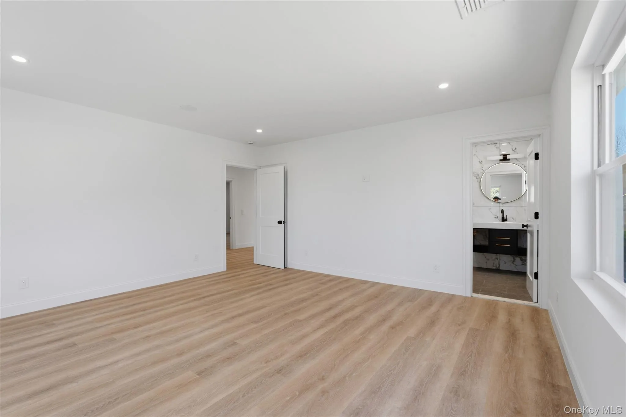Spacious room featuring light wood-finish flooring, recessed lighting, and a private bathroom with a dark vanity, round mirror, and black fixtures Spacious room featuring light wood-finish flooring, recessed lighting, and a private bathroom with a dark vanity, round mirror, and black fixtures