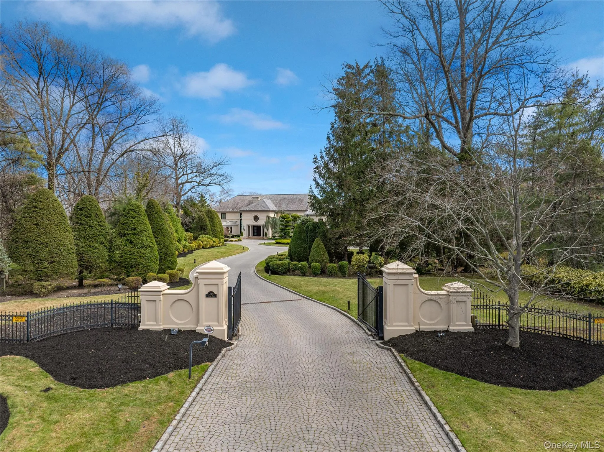 Gated entrance with paver driveway, manicured landscaping, and mature trees framing the property Gated entrance with paver driveway, manicured landscaping, and mature trees framing the property