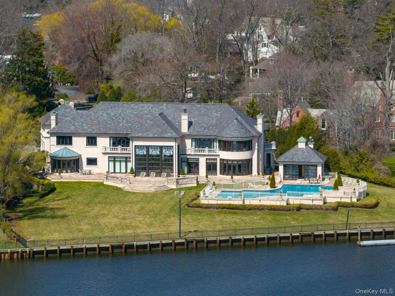 Waterfront property featuring a stucco exterior, slate roof, expansive rear patio, in-ground swimming pool, and a dedicated pool house Waterfront property featuring a stucco exterior, slate roof, expansive rear patio, in-ground swimming pool, and a dedicated pool house