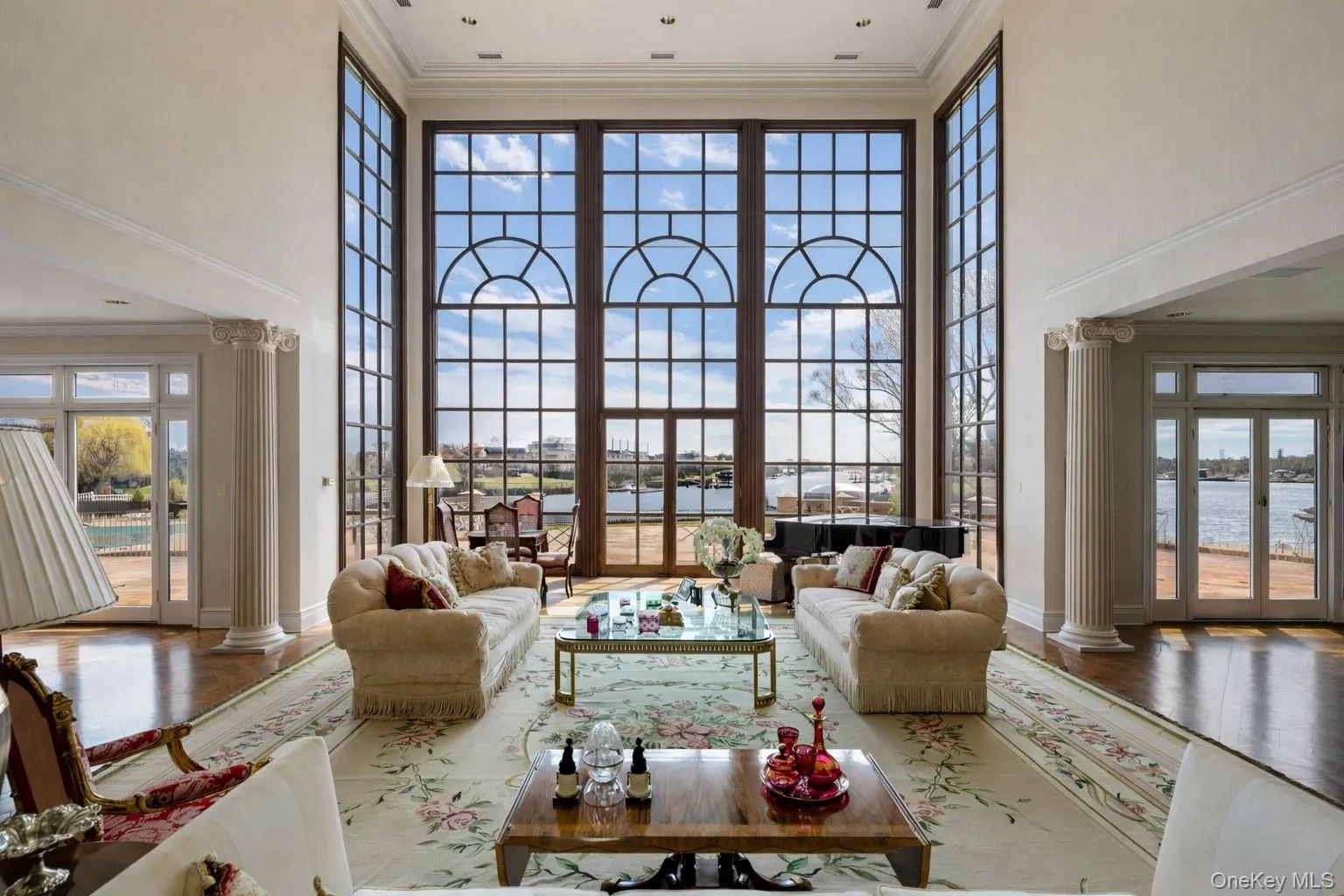 Grand living space featuring floor-to-ceiling windows with arched transoms, extensive architectural columns, and wood-finish flooring Grand living space featuring floor-to-ceiling windows with arched transoms, extensive architectural columns, and wood-finish flooring