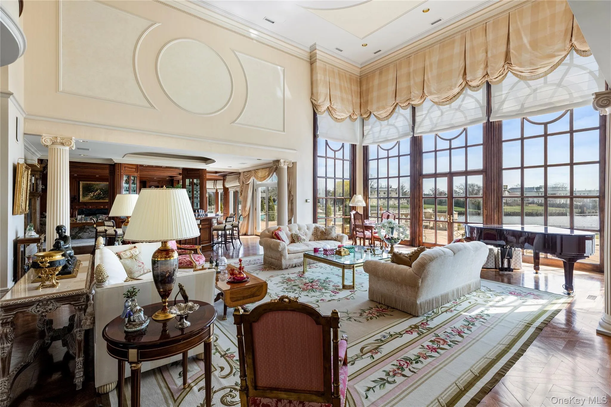 Grand living space featuring a two-story ceiling with architectural paneling, floor-to-ceiling windows, wood-finish flooring, and white columns Grand living space featuring a two-story ceiling with architectural paneling, floor-to-ceiling windows, wood-finish flooring, and white columns