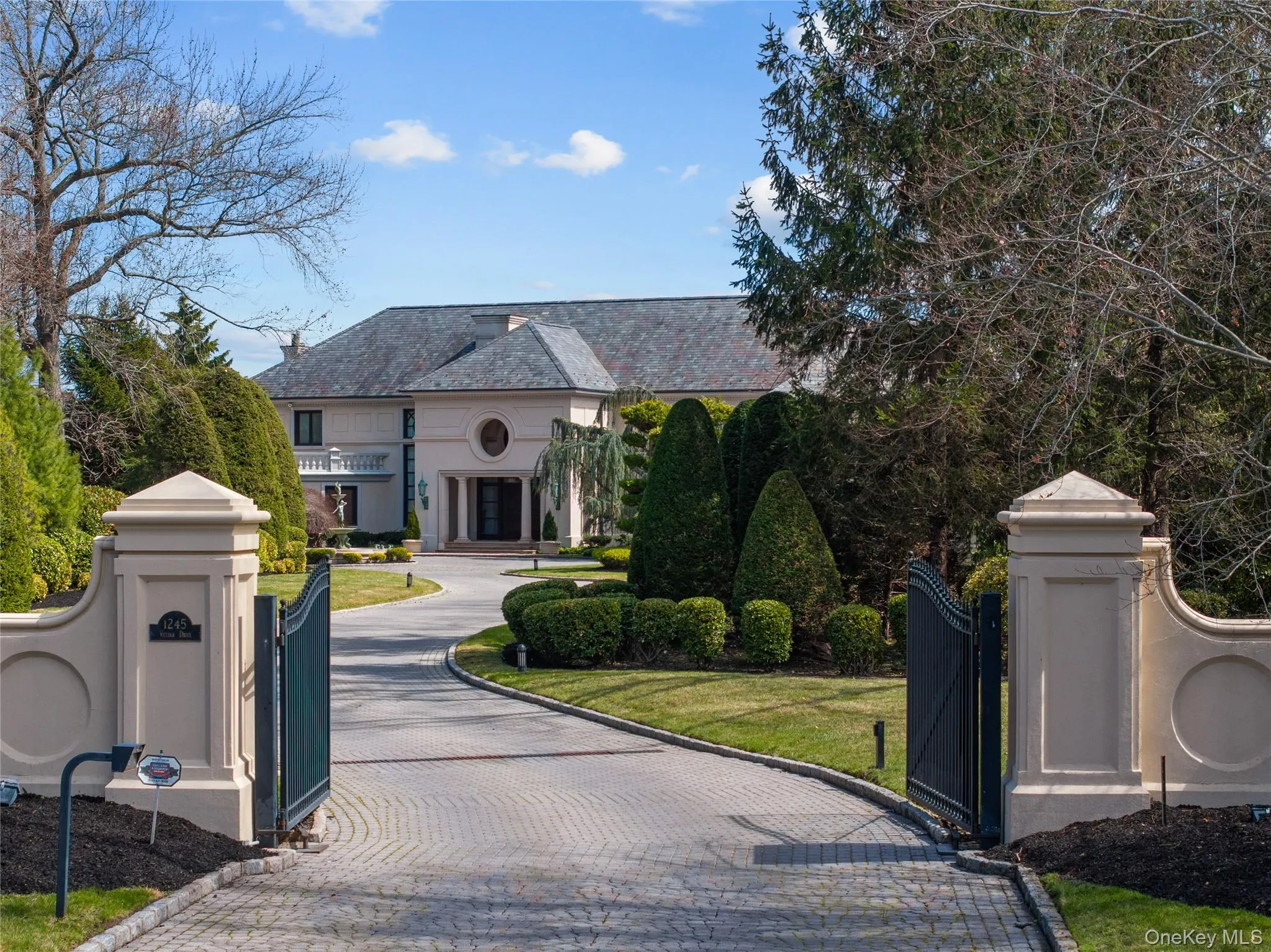 Gated entry featuring a paved driveway, manicured lawns, mature landscaping, and a stucco exterior residence with a slate-style roof Gated entry featuring a paved driveway, manicured lawns, mature landscaping, and a stucco exterior residence with a slate-style roof