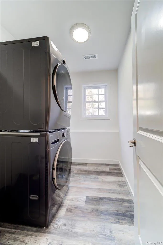 Dedicated laundry area featuring stacked front-load machines, wood-finish flooring, a single window, white walls, and an overhead light fixture Dedicated laundry area featuring stacked front-load machines, wood-finish flooring, a single window, white walls, and an overhead light fixture