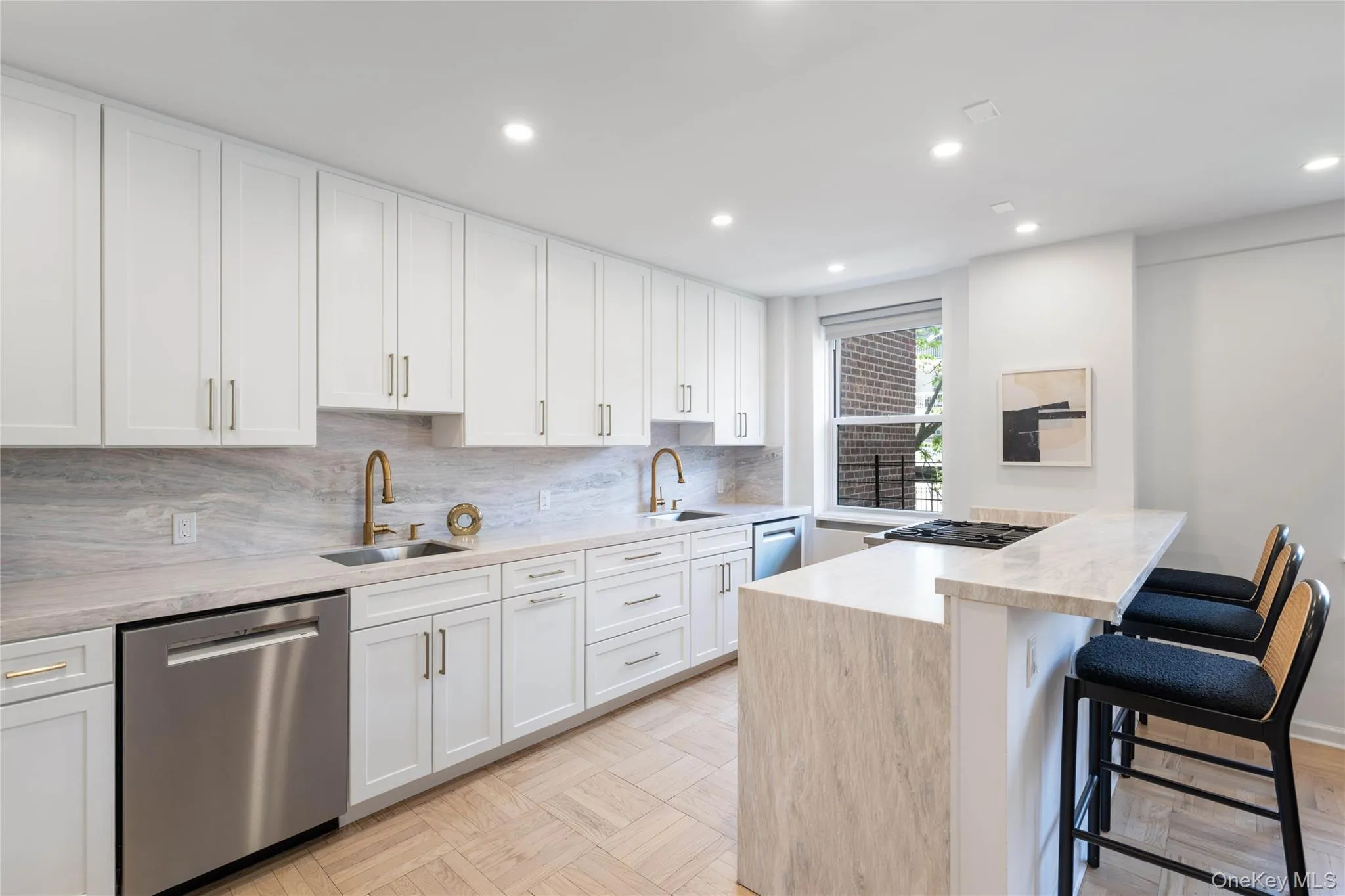 Gourmet kitchen featuring extensive white cabinetry, light-toned stone countertops with matching backsplash, stainless steel appliances, and a central island with a breakfast bar and waterfall edge. Gourmet kitchen featuring extensive white cabinetry, light-toned stone countertops with matching backsplash, stainless steel appliances, and a central island with a breakfast bar and waterfall edge.