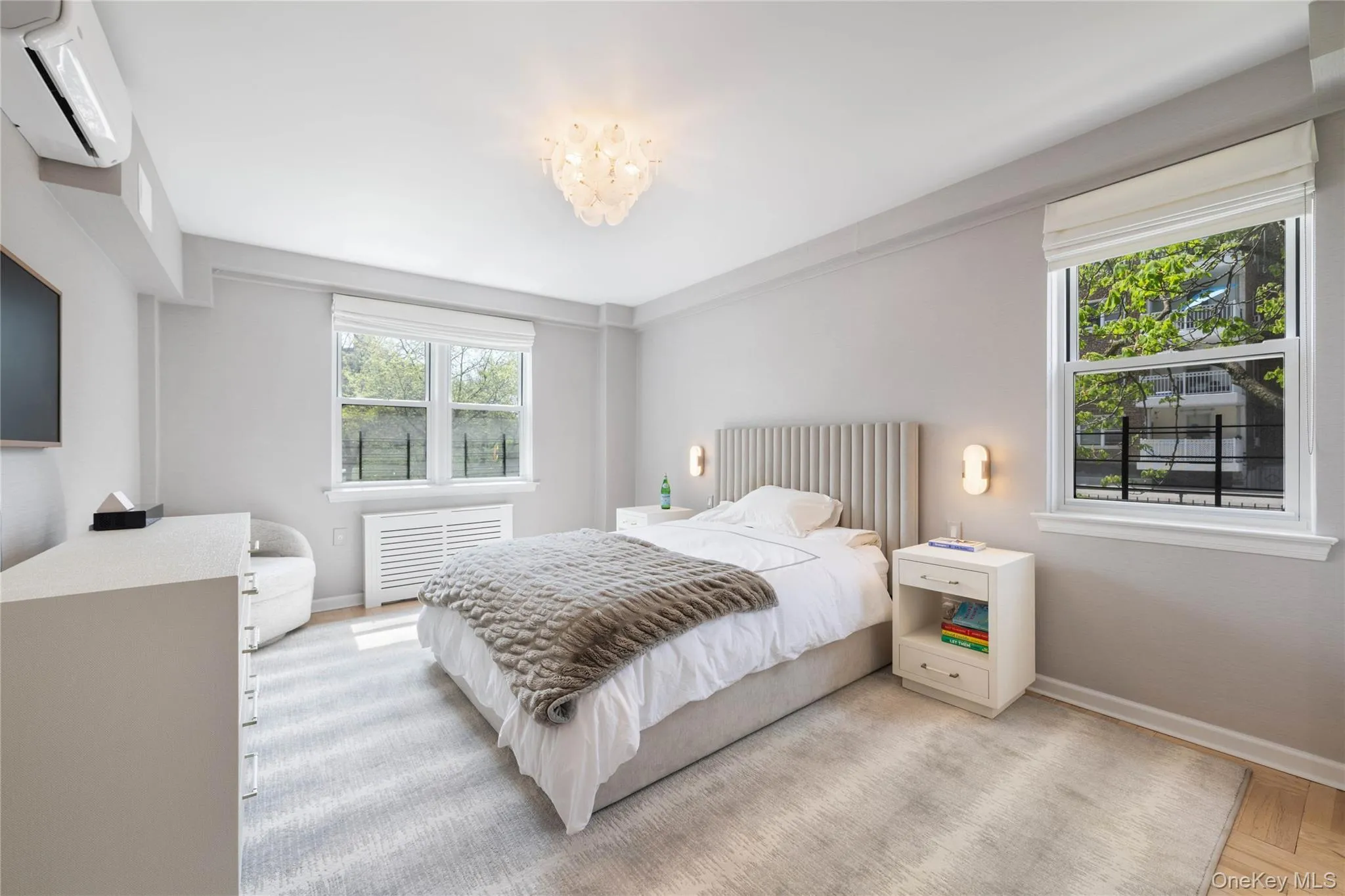 Light-filled bedroom featuring two double-hung windows with Roman shades, a decorative ceiling fixture, and two wall-mounted sconces Light-filled bedroom featuring two double-hung windows with Roman shades, a decorative ceiling fixture, and two wall-mounted sconces