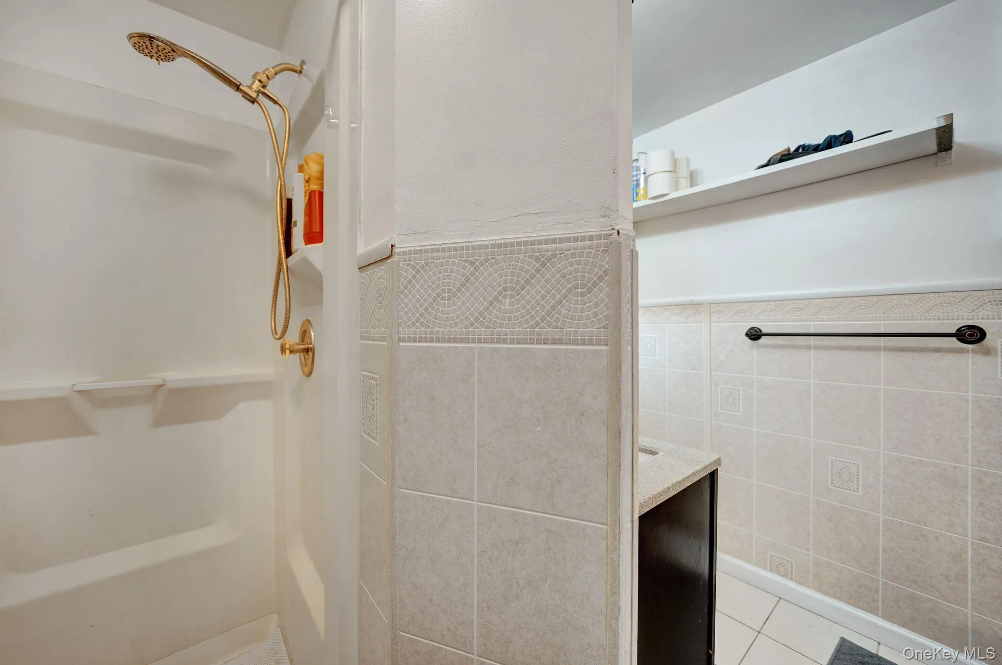 Bathroom featuring a white shower stall with a gold-toned shower head, tiled walls with decorative accents, a dark vanity cabinet with a light-colored countertop, and a wall-mounted shelf Bathroom featuring a white shower stall with a gold-toned shower head, tiled walls with decorative accents, a dark vanity cabinet with a light-colored countertop, and a wall-mounted shelf