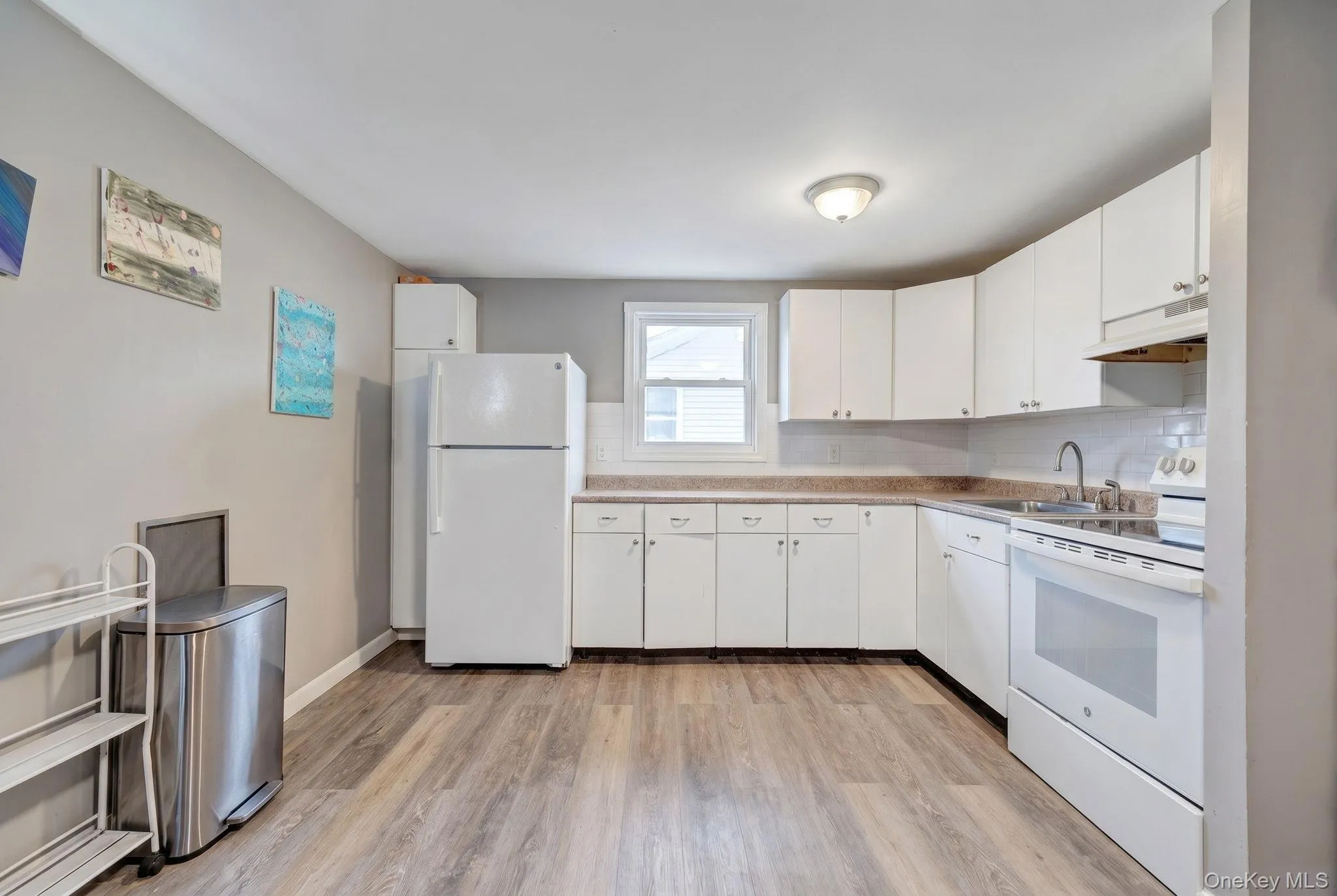 Kitchen featuring wood-finish flooring, white cabinetry, laminate countertops, a white subway tile backsplash, and a white appliance suite Kitchen featuring wood-finish flooring, white cabinetry, laminate countertops, a white subway tile backsplash, and a white appliance suite