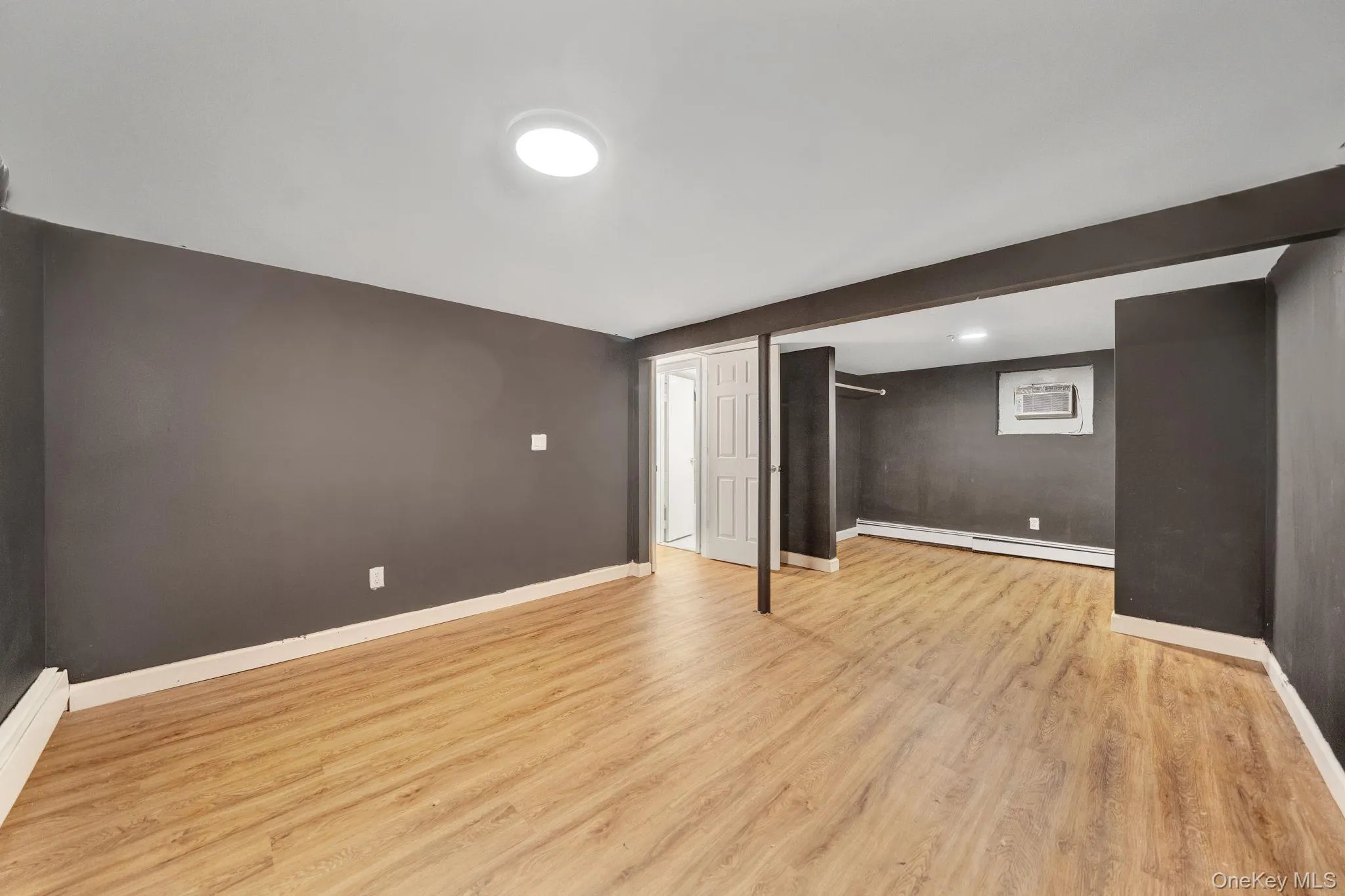 Spacious room featuring wood-finish flooring, dark wall paint, white baseboards, and a circular ceiling light fixture basement Spacious room featuring wood-finish flooring, dark wall paint, white baseboards, and a circular ceiling light fixture basement