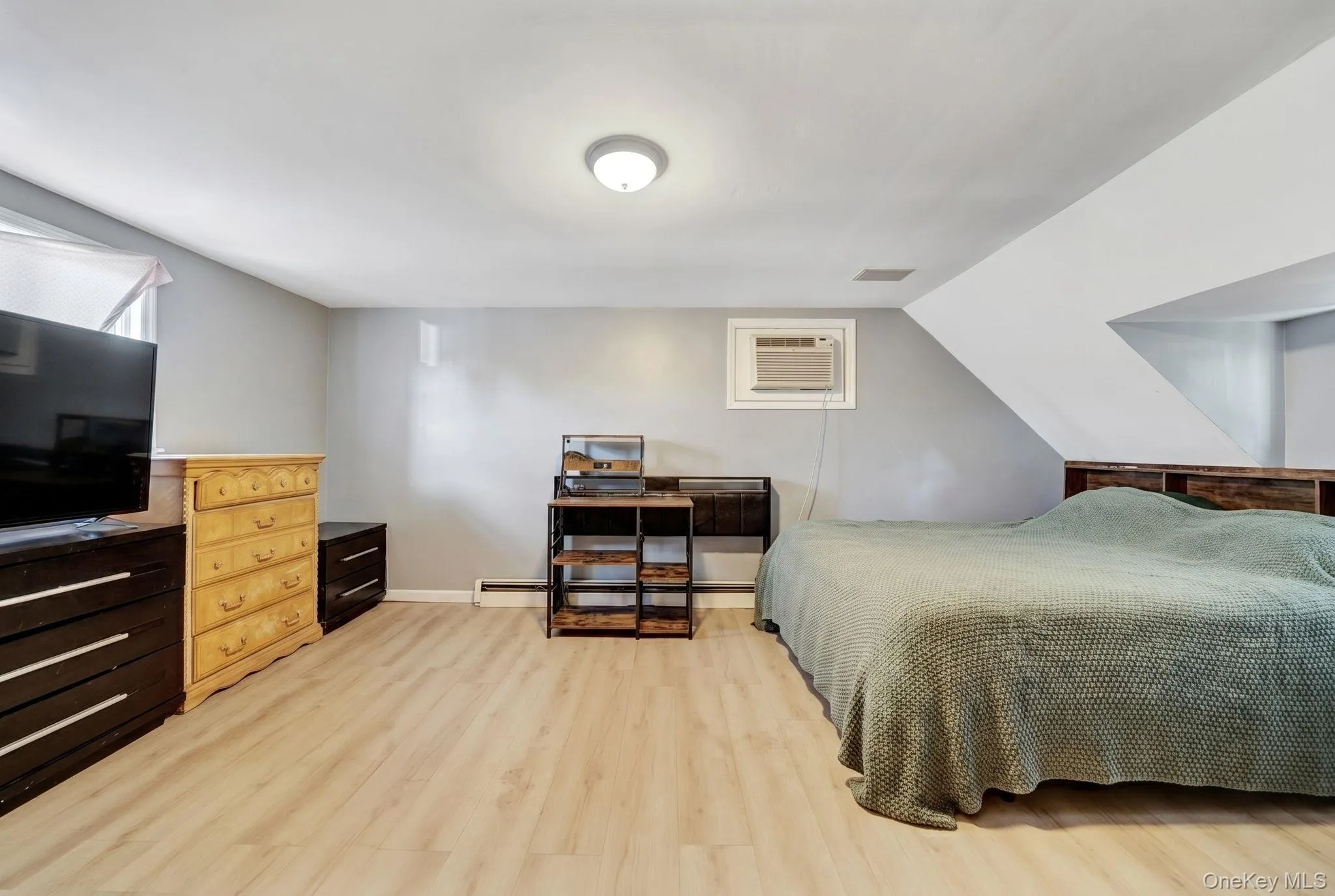 Spacious room featuring light wood-finish flooring, a window-mounted air conditioning unit, and a built-in wooden headboard with shelving Spacious room featuring light wood-finish flooring, a window-mounted air conditioning unit, and a built-in wooden headboard with shelving