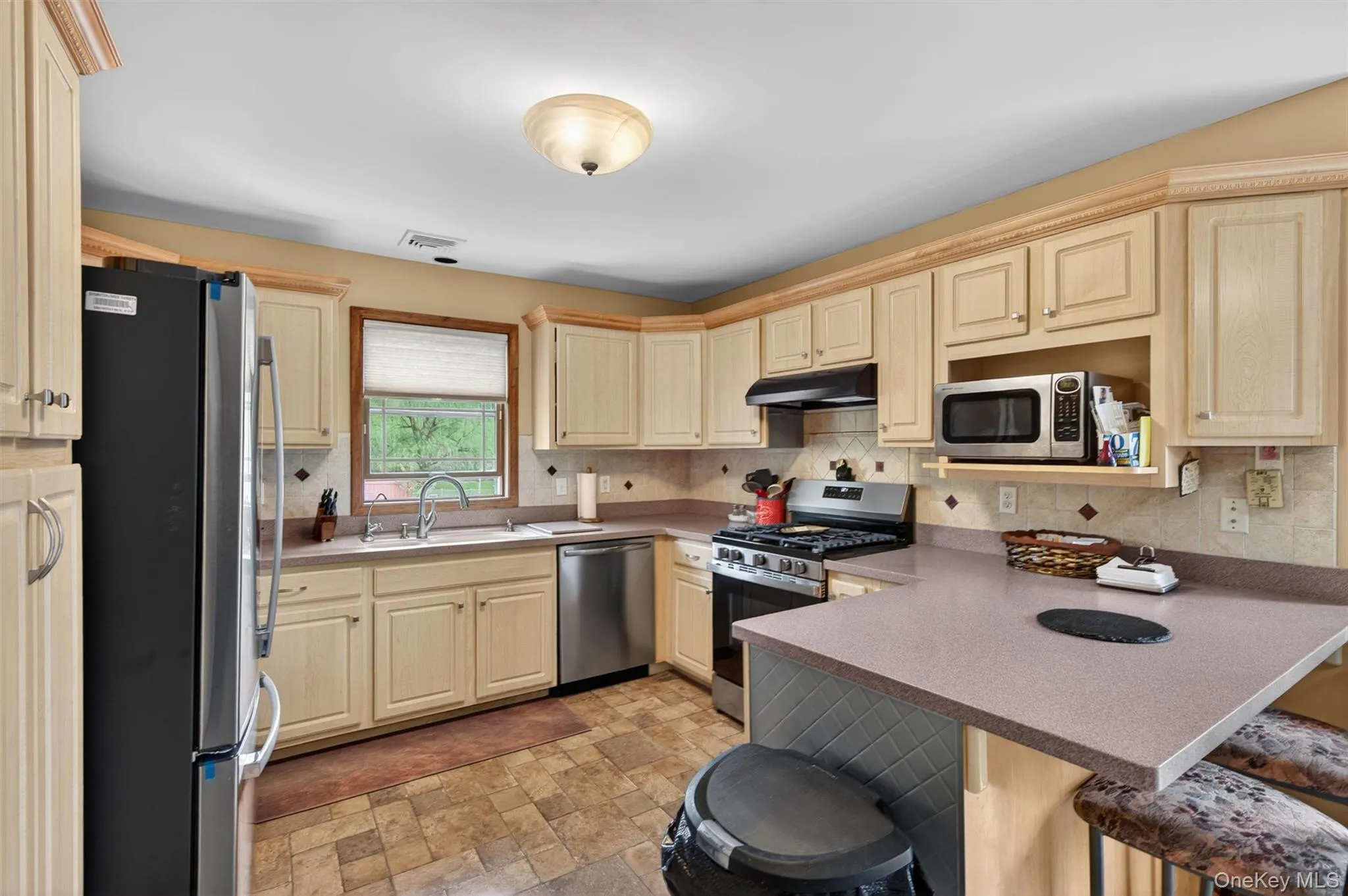Kitchen featuring light-toned cabinetry, patterned tile flooring, an integrated breakfast bar, stainless steel appliances, and a window with a garden outlook Kitchen featuring light-toned cabinetry, patterned tile flooring, an integrated breakfast bar, stainless steel appliances, and a window with a garden outlook