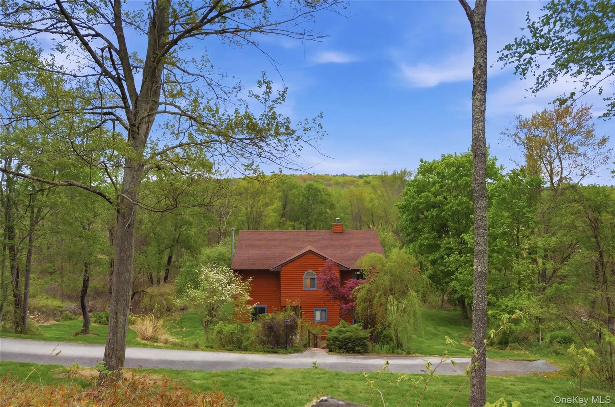Red-toned contemporary house with a gabled roof set amidst mature trees and lush greenery with a driveway leading down to the home. Red-toned contemporary house with a gabled roof set amidst mature trees and lush greenery with a driveway leading down to the home.