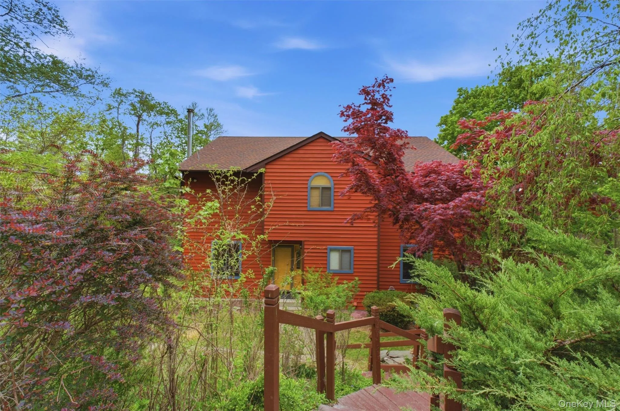 Red-toned contemporary house with a gabled roof set amidst mature trees and lush greenery with a staircase leading down to the home. Red-toned contemporary house with a gabled roof set amidst mature trees and lush greenery with a staircase leading down to the home.