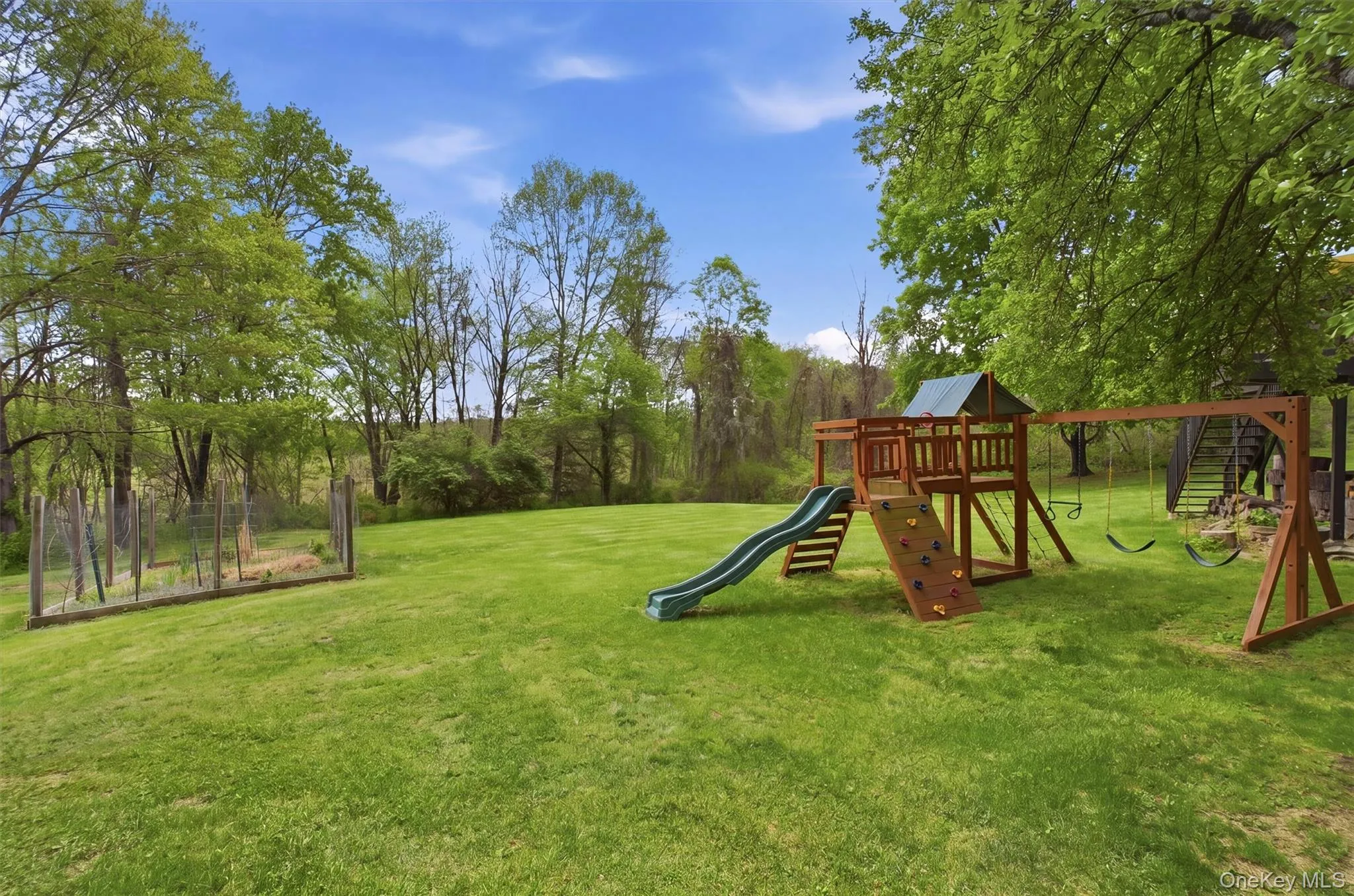 Expansive green lawn featuring a wooden playset with slide and swings, surrounded by mature trees and privacy. Expansive green lawn featuring a wooden playset with slide and swings, surrounded by mature trees and privacy.
