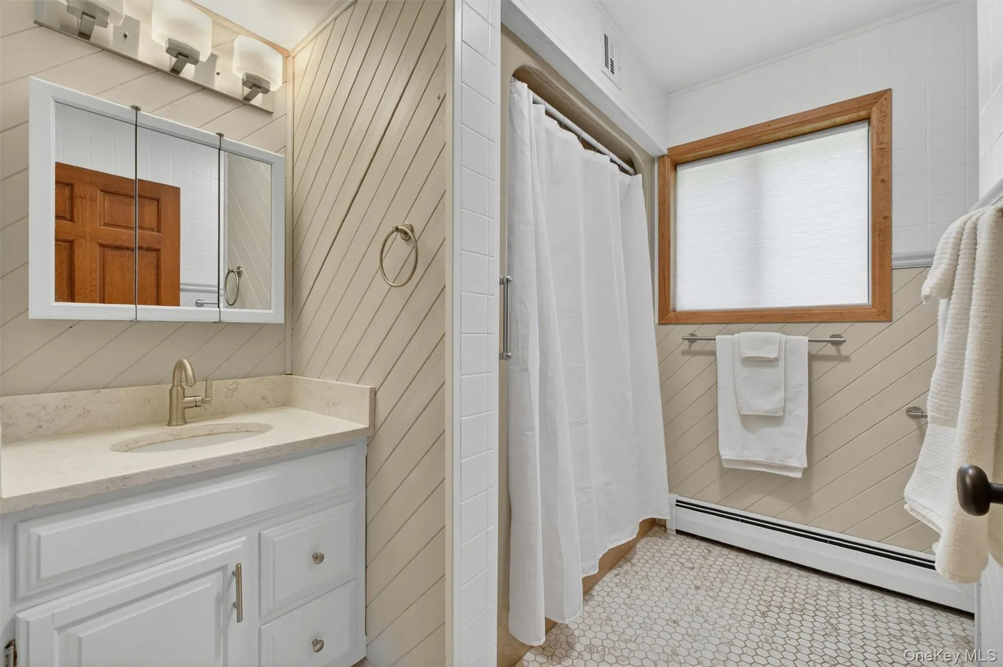 Neutral toned bathroom featuring a white vanity with a light-toned countertop, a recessed medicine cabinet with overhead lighting, diagonal paneling on the walls, a shower enclosure, and hexagon tile flooring. Neutral toned bathroom featuring a white vanity with a light-toned countertop, a recessed medicine cabinet with overhead lighting, diagonal paneling on the walls, a shower enclosure, and hexagon tile flooring.