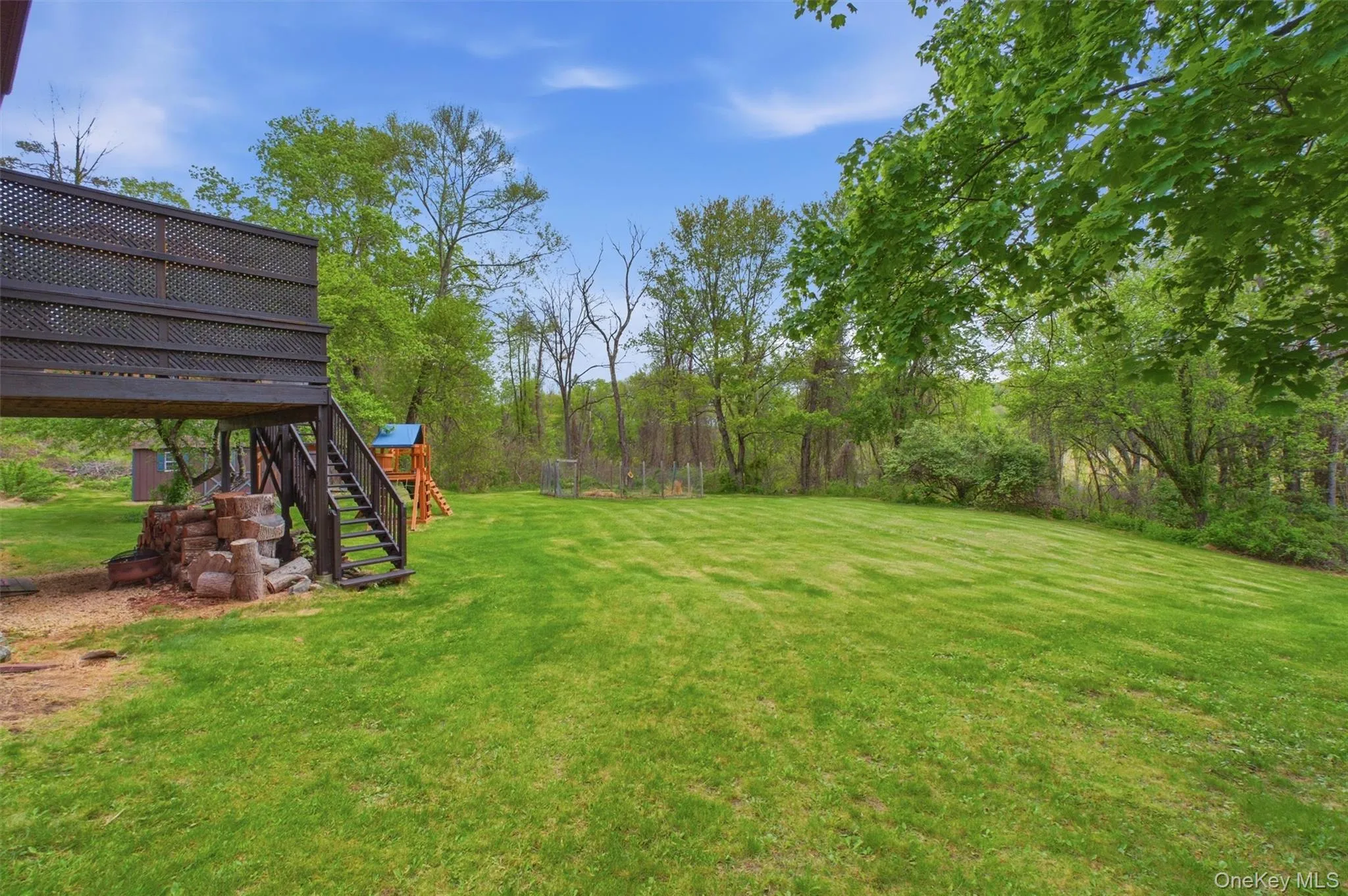 Expansive green lawn with mature perimeter trees, featuring a raised deck with lattice screening and stairs, and a partial privacy fence. Expansive green lawn with mature perimeter trees, featuring a raised deck with lattice screening and stairs, and a partial privacy fence.