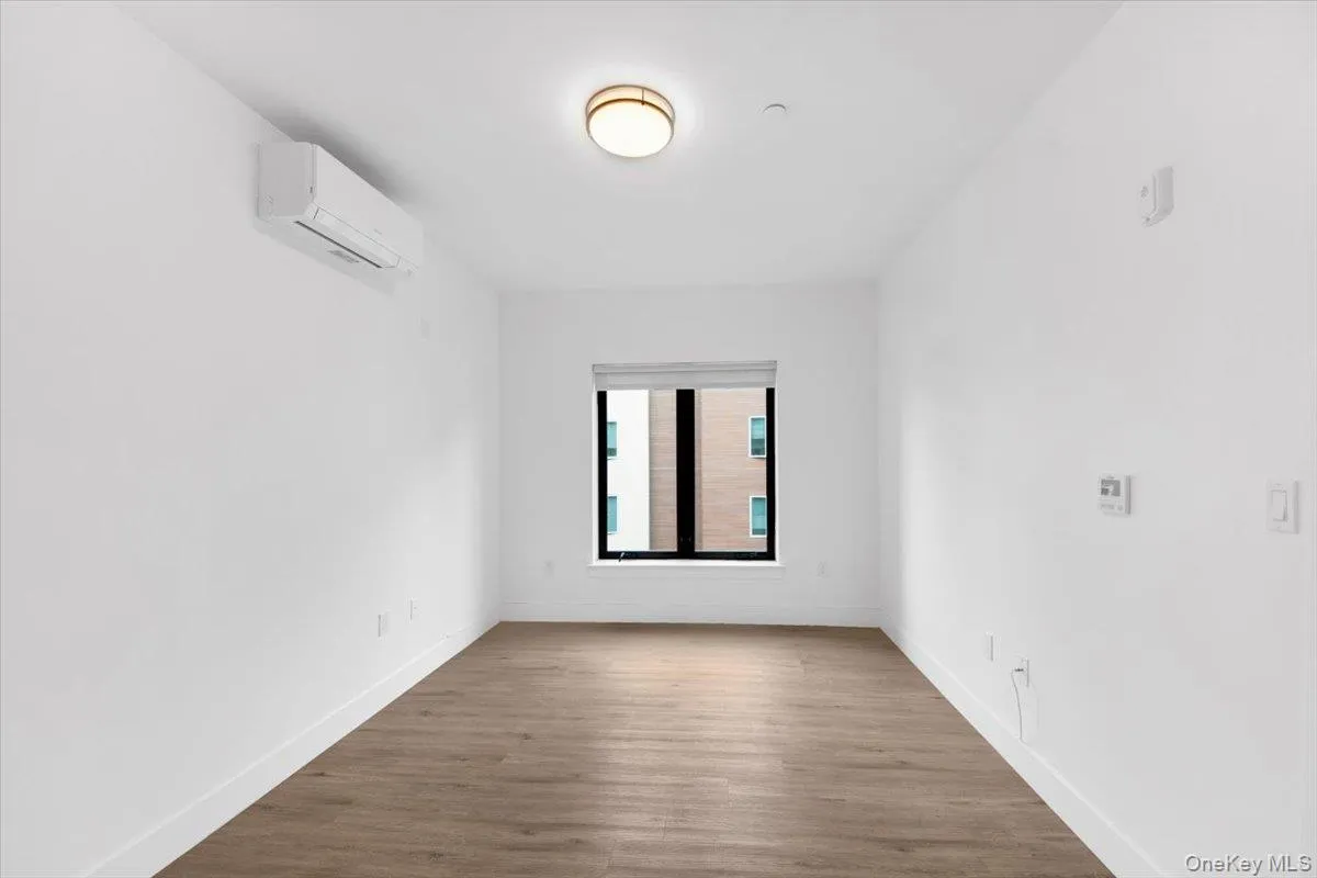 Bright interior space featuring wood-finish flooring, white walls, and a window with a roller shade Bright interior space featuring wood-finish flooring, white walls, and a window with a roller shade
