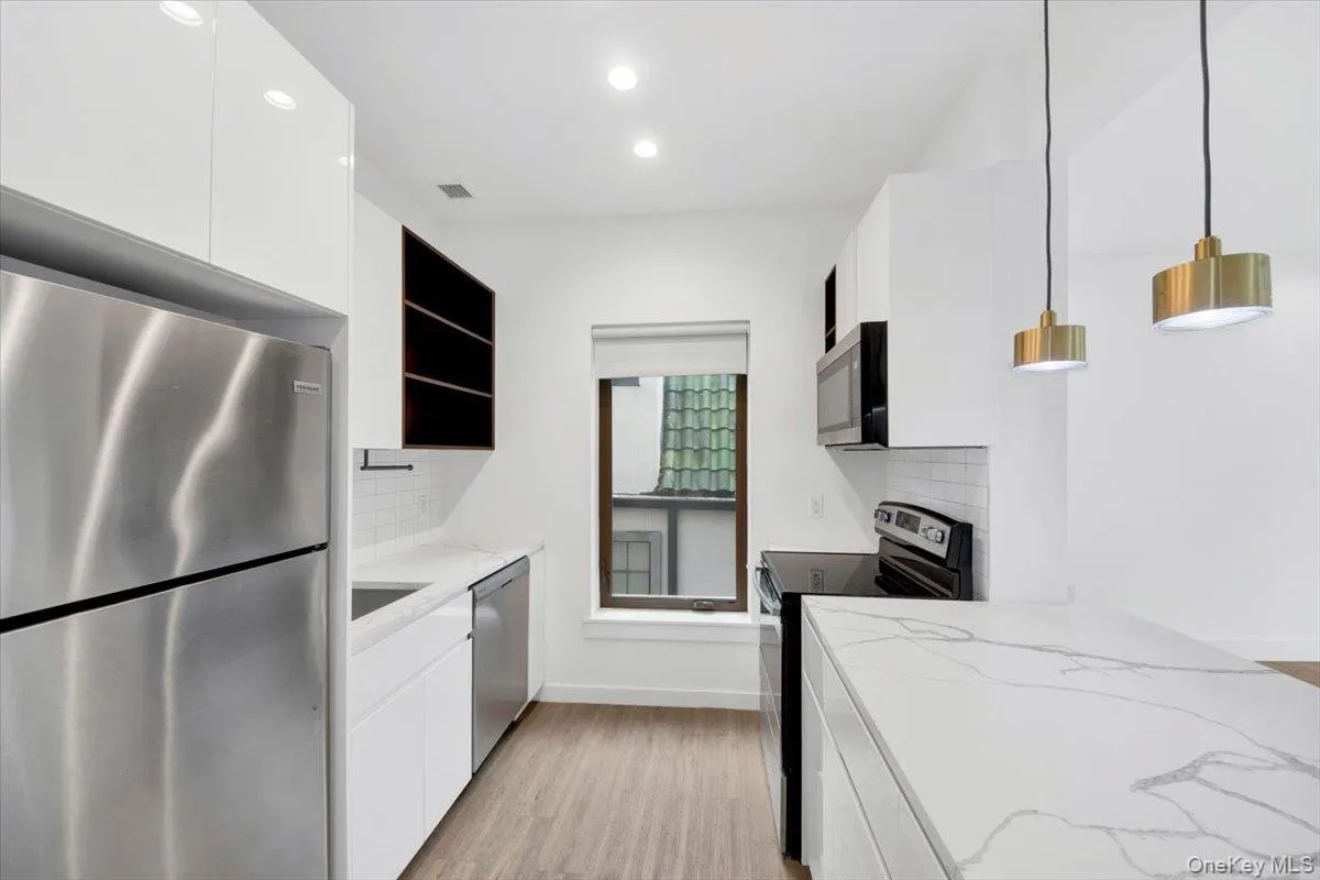Galley kitchen featuring stainless steel appliances, white flat-panel cabinetry, white subway tile backsplash, white countertops with veining, and wood-finish flooring Galley kitchen featuring stainless steel appliances, white flat-panel cabinetry, white subway tile backsplash, white countertops with veining, and wood-finish flooring