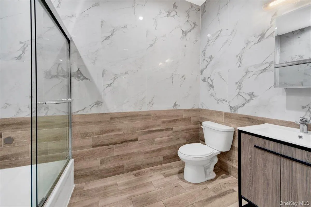 Bathroom featuring a glass shower enclosure over a tub, wood-finish flooring, half-wall wood-look tiling, full-wall marble-look tiling, and a vanity with an integrated sink Bathroom featuring a glass shower enclosure over a tub, wood-finish flooring, half-wall wood-look tiling, full-wall marble-look tiling, and a vanity with an integrated sink