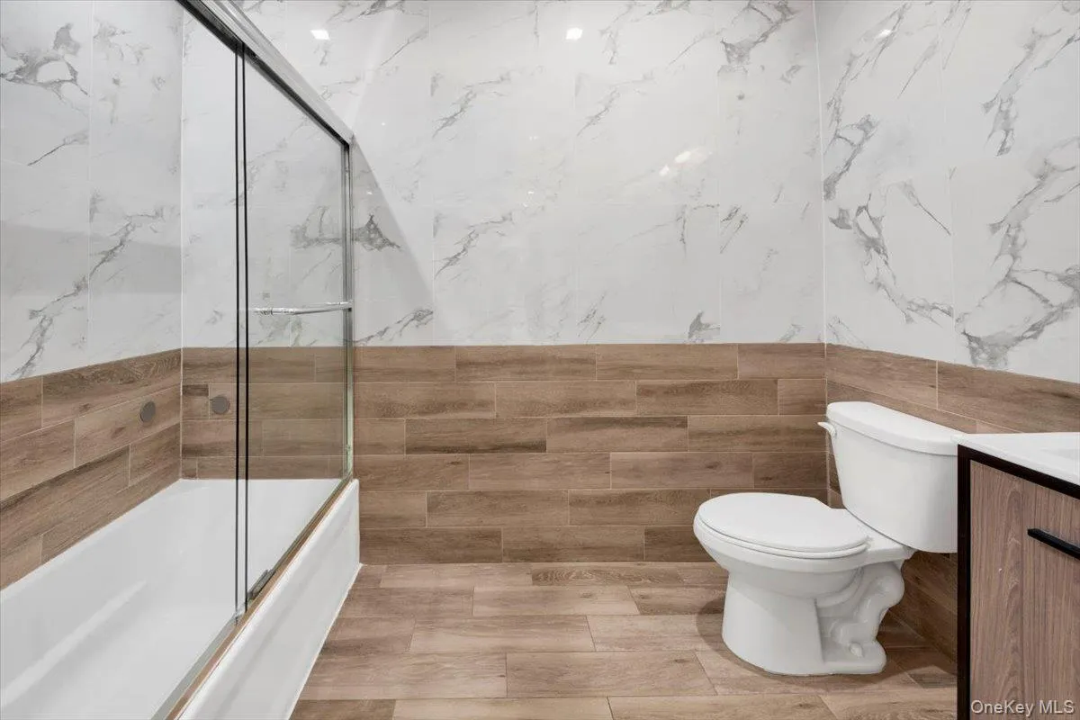 Bathroom featuring a full tub with sliding glass doors, white toilet, and wood-finish floor and wall tiling complemented by white marble-look wall tiling Bathroom featuring a full tub with sliding glass doors, white toilet, and wood-finish floor and wall tiling complemented by white marble-look wall tiling