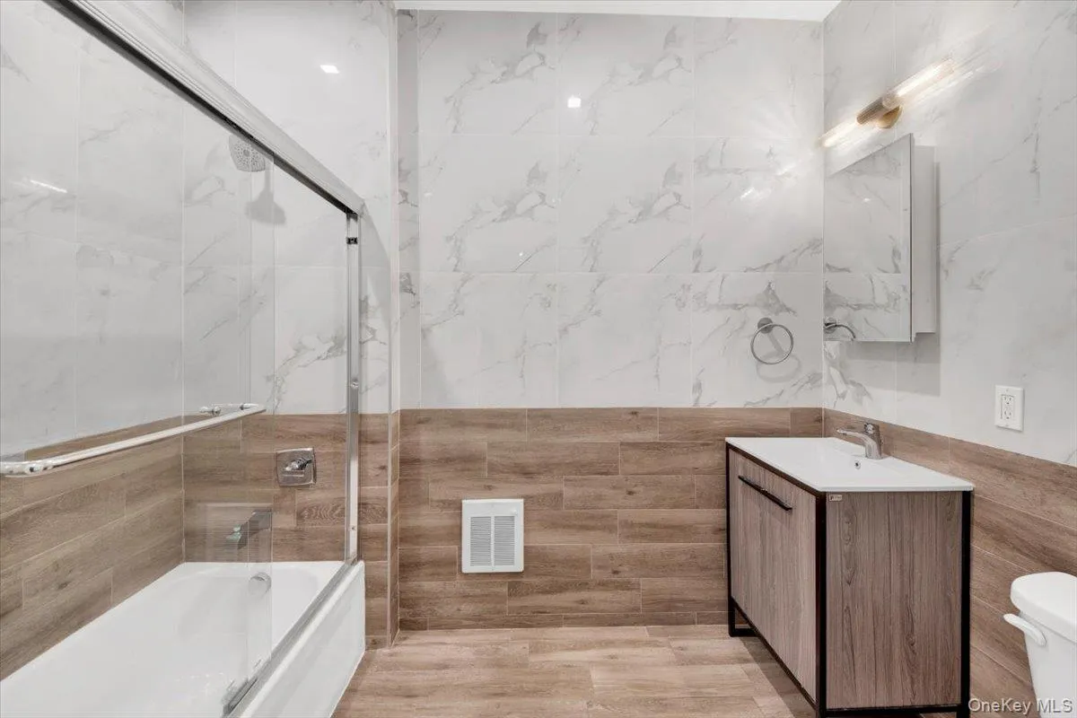 Bathroom featuring a full tub with sliding glass shower doors, wood-finish tile flooring, and contrasting white marble-patterned wall tiles Bathroom featuring a full tub with sliding glass shower doors, wood-finish tile flooring, and contrasting white marble-patterned wall tiles