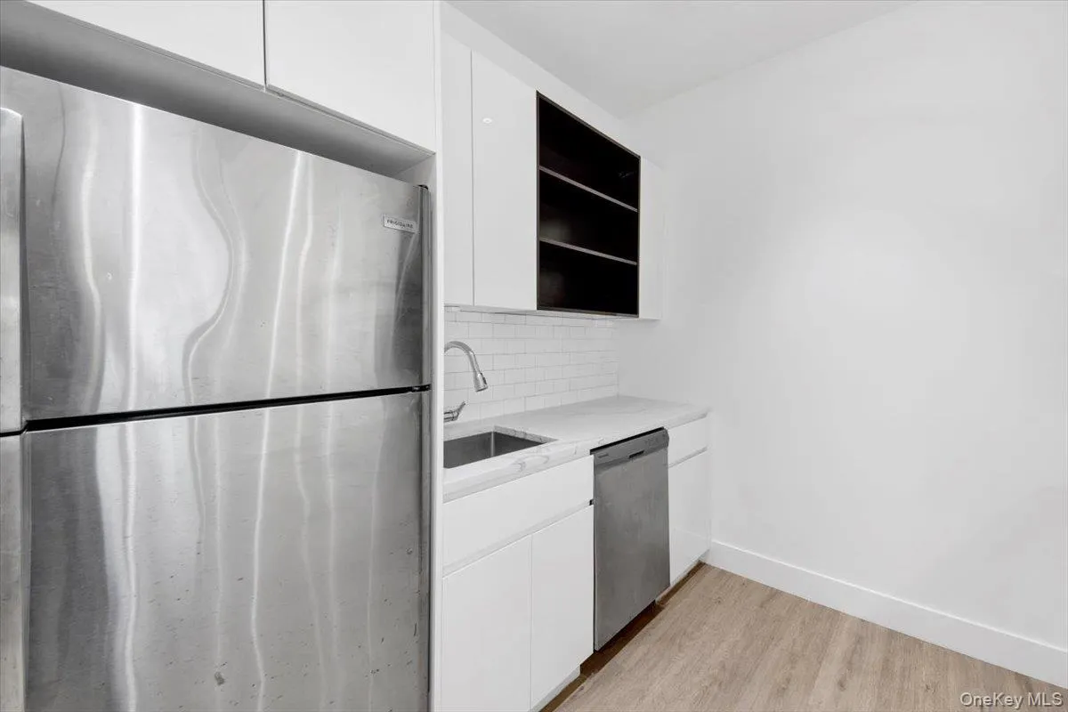 Kitchenette featuring a stainless steel refrigerator, white flat-panel cabinetry, white subway tile backsplash, white countertops, a stainless steel sink with a gooseneck faucet, and wood-finish flooring Kitchenette featuring a stainless steel refrigerator, white flat-panel cabinetry, white subway tile backsplash, white countertops, a stainless steel sink with a gooseneck faucet, and wood-finish flooring