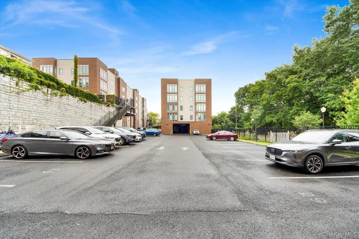 Paved parking area featuring designated spaces, surrounding modern multi-story buildings with brick and light-toned facades, and a retaining wall with greenery Paved parking area featuring designated spaces, surrounding modern multi-story buildings with brick and light-toned facades, and a retaining wall with greenery