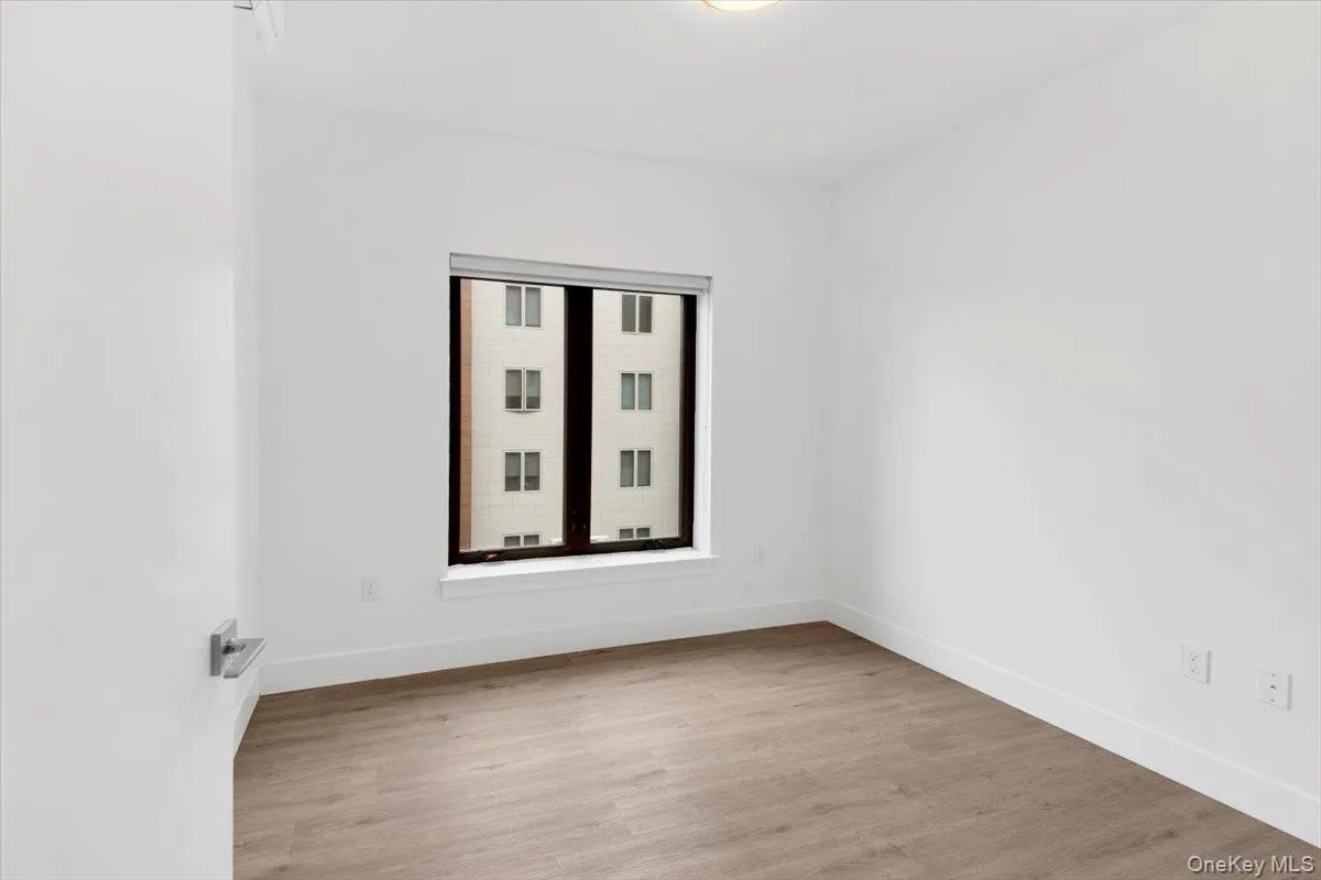 Bright interior space featuring a window with a roller shade, wood-finish flooring, white walls, and white baseboards Bright interior space featuring a window with a roller shade, wood-finish flooring, white walls, and white baseboards