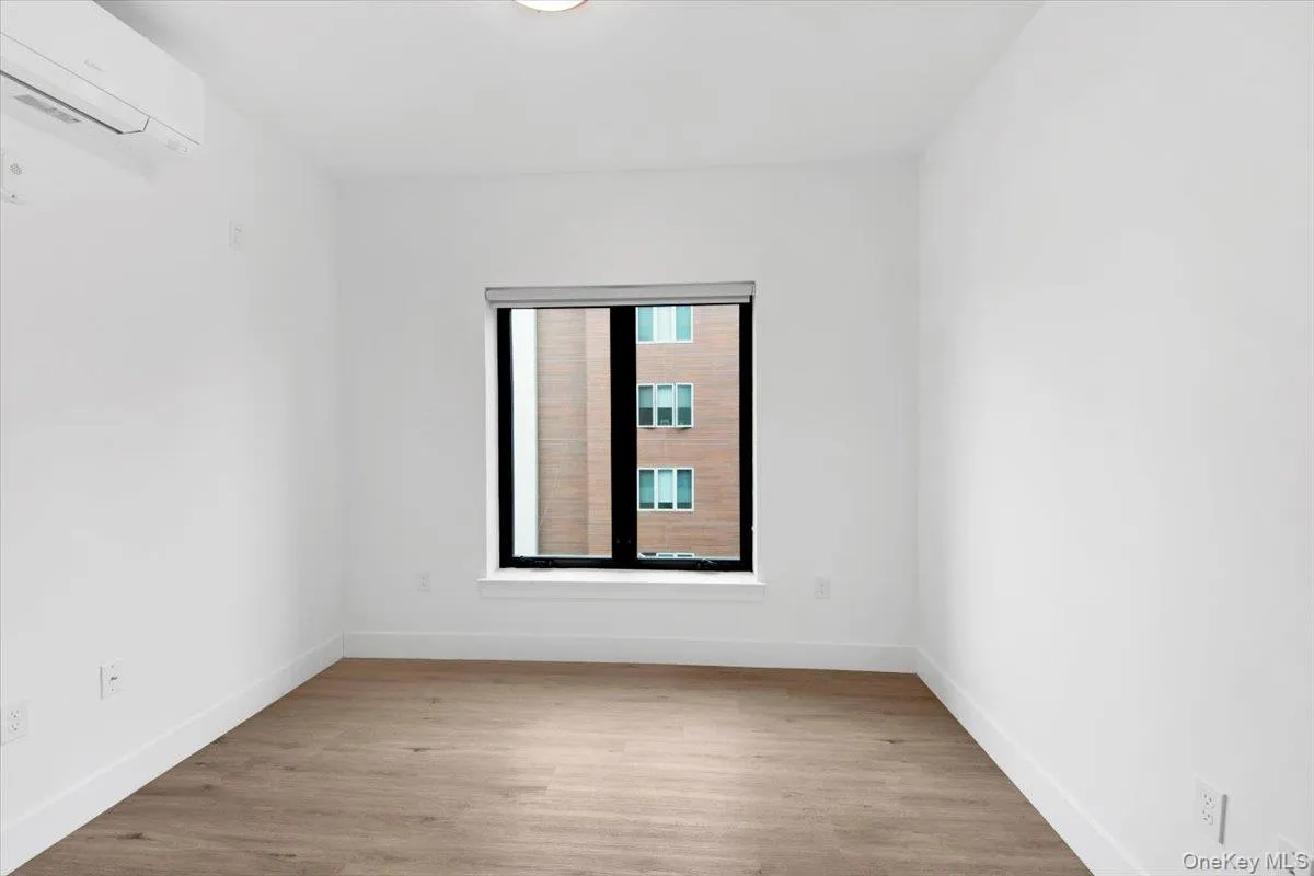 Room featuring wood-finish flooring, a contemporary window with a dark frame, white walls, and a ceiling-mounted light fixture Room featuring wood-finish flooring, a contemporary window with a dark frame, white walls, and a ceiling-mounted light fixture