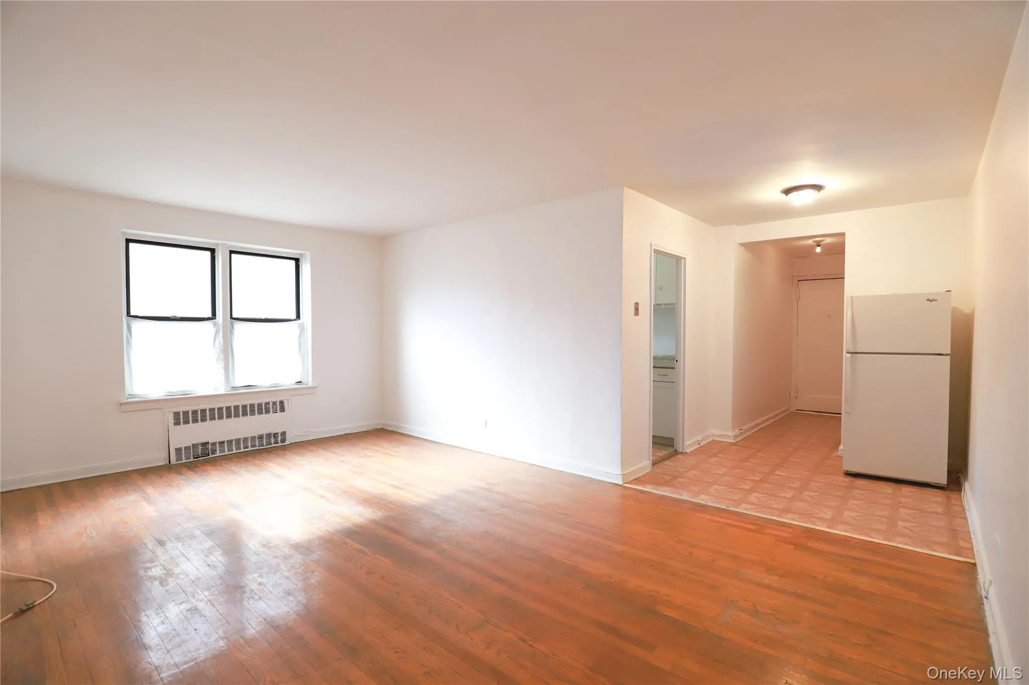Spacious room featuring wood-finish flooring, white walls, and a large window with a radiator below Spacious room featuring wood-finish flooring, white walls, and a large window with a radiator below