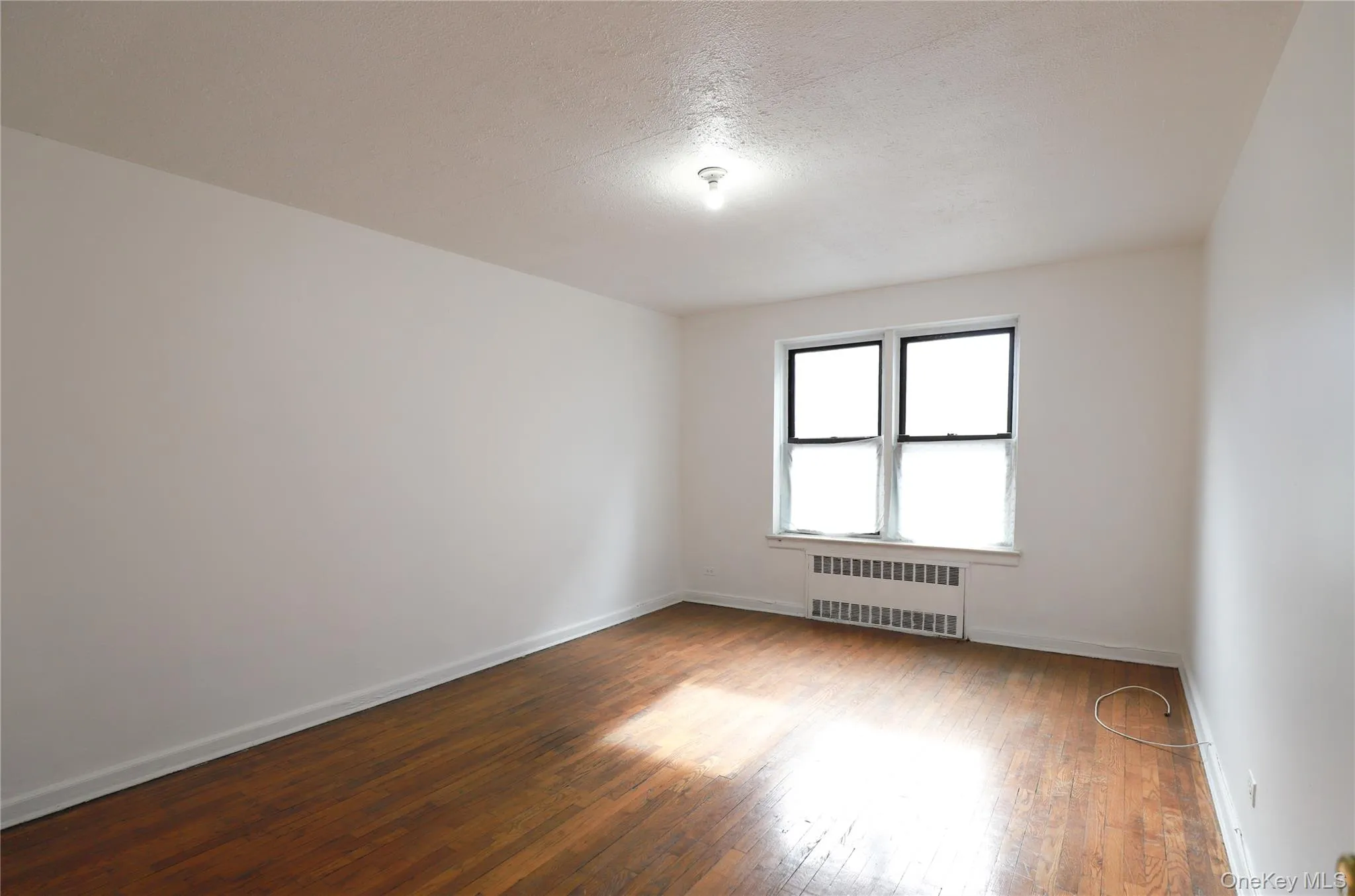 Hardwood flooring throughout featuring white baseboards, a window with a radiator below, and a flush-mount ceiling light fixture Hardwood flooring throughout featuring white baseboards, a window with a radiator below, and a flush-mount ceiling light fixture