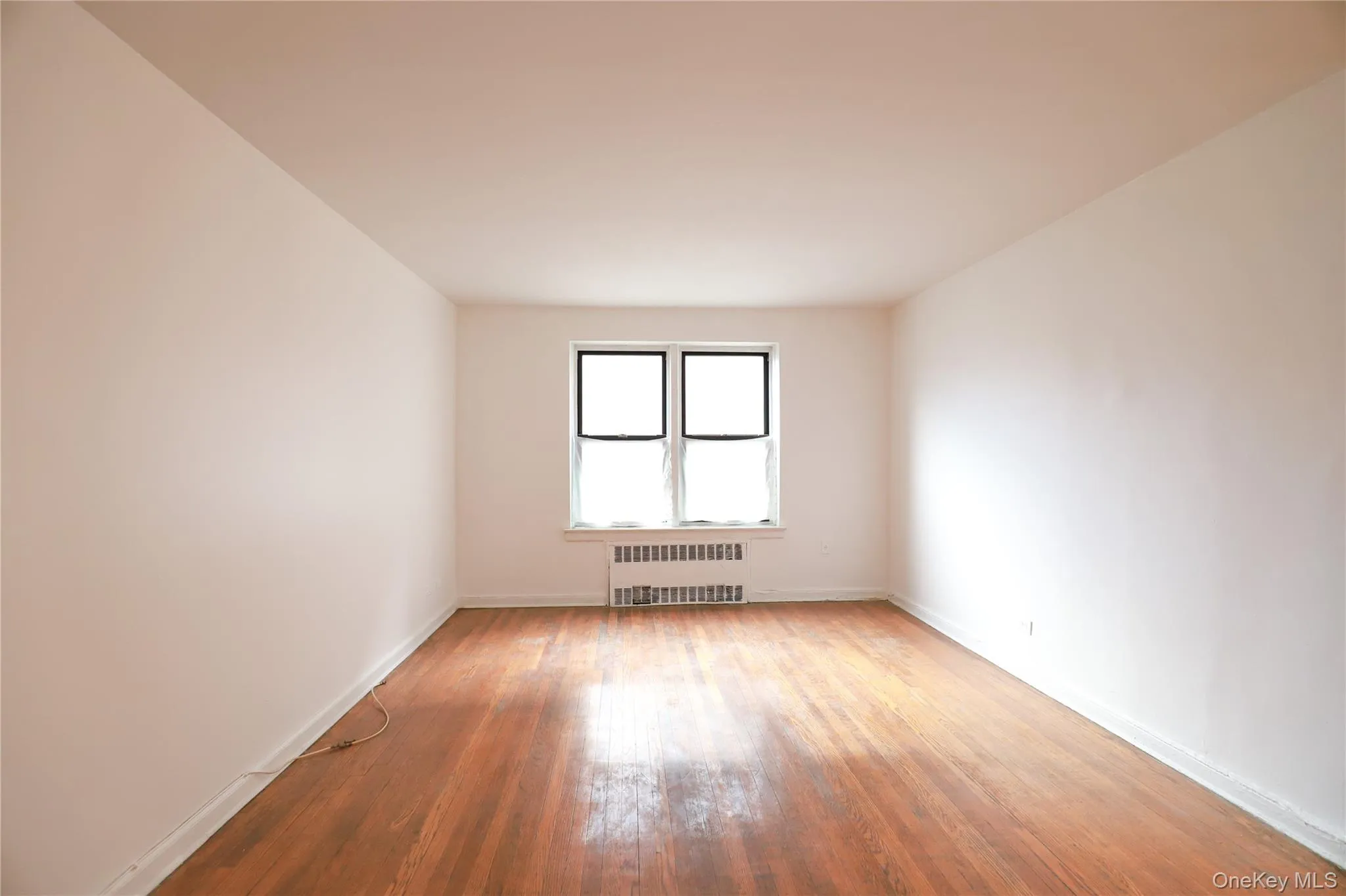 Spacious interior featuring wood-finish flooring, white walls, and a window with a radiator below Spacious interior featuring wood-finish flooring, white walls, and a window with a radiator below