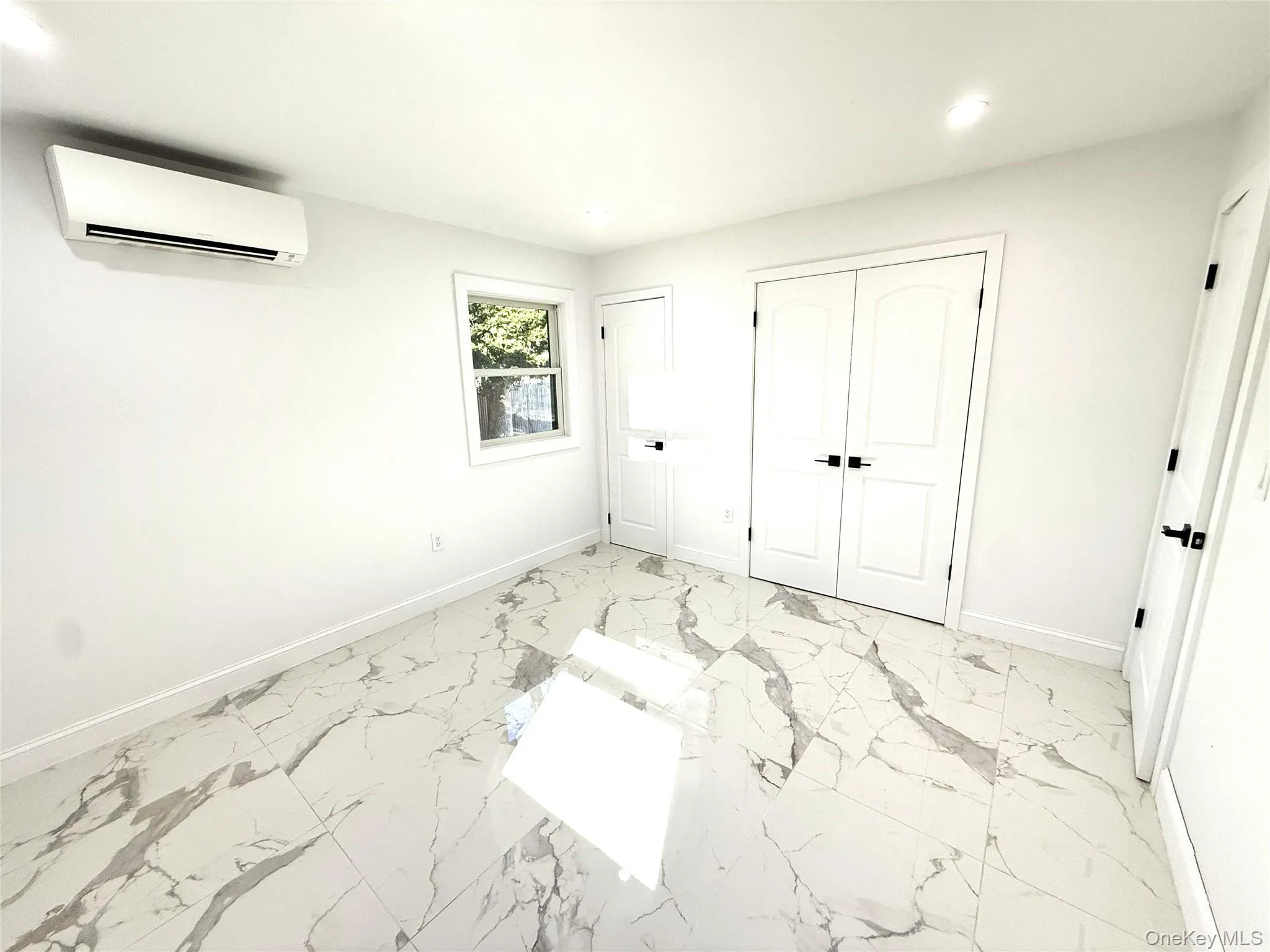 Spacious room featuring white walls, large format marble-look tile flooring, and a mini-split air conditioning unit Spacious room featuring white walls, large format marble-look tile flooring, and a mini-split air conditioning unit