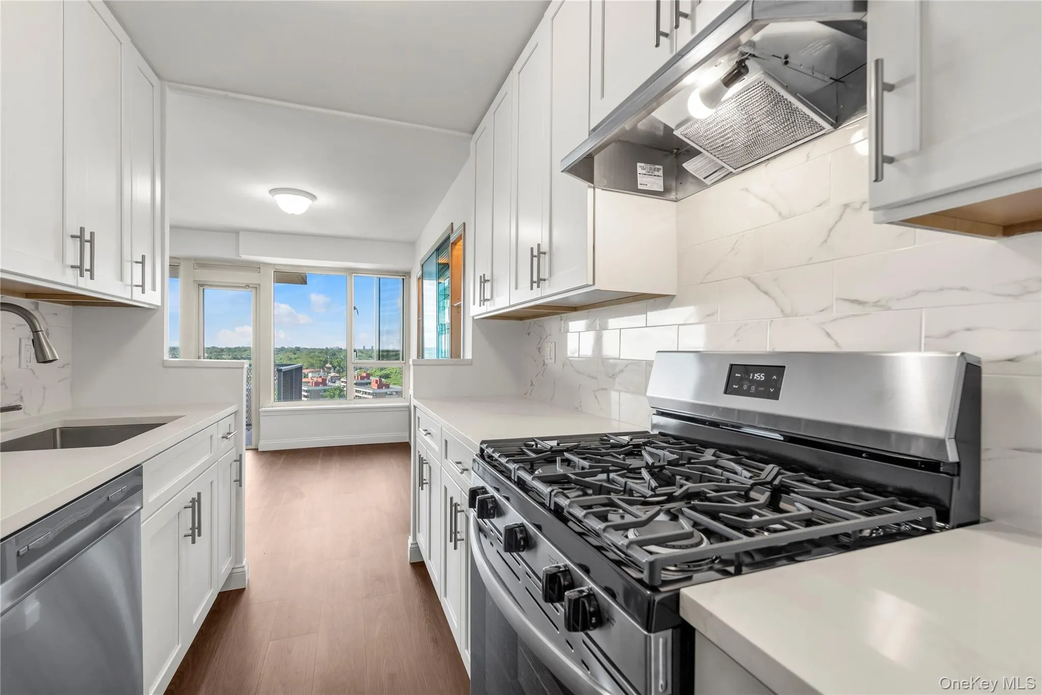 NEW Galley kitchen featuring white shaker cabinetry, white countertops, a tiled backsplash, wood-finish flooring, and stainless steel appliances NEW Galley kitchen featuring white shaker cabinetry, white countertops, a tiled backsplash, wood-finish flooring, and stainless steel appliances