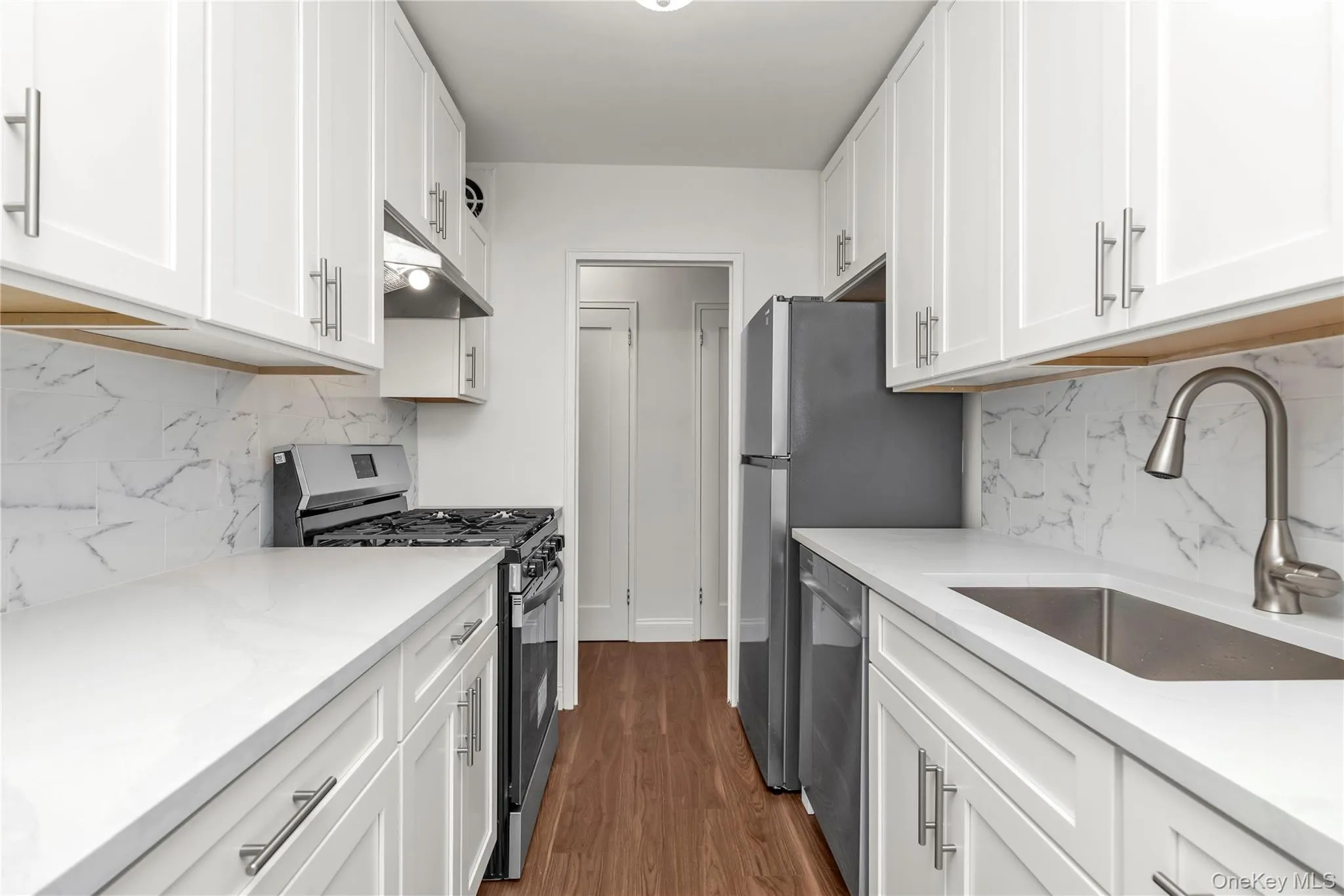 NEW Galley kitchen featuring white shaker cabinetry, white countertops, a tiled backsplash, wood-finish flooring, and stainless steel appliances NEW Galley kitchen featuring white shaker cabinetry, white countertops, a tiled backsplash, wood-finish flooring, and stainless steel appliances