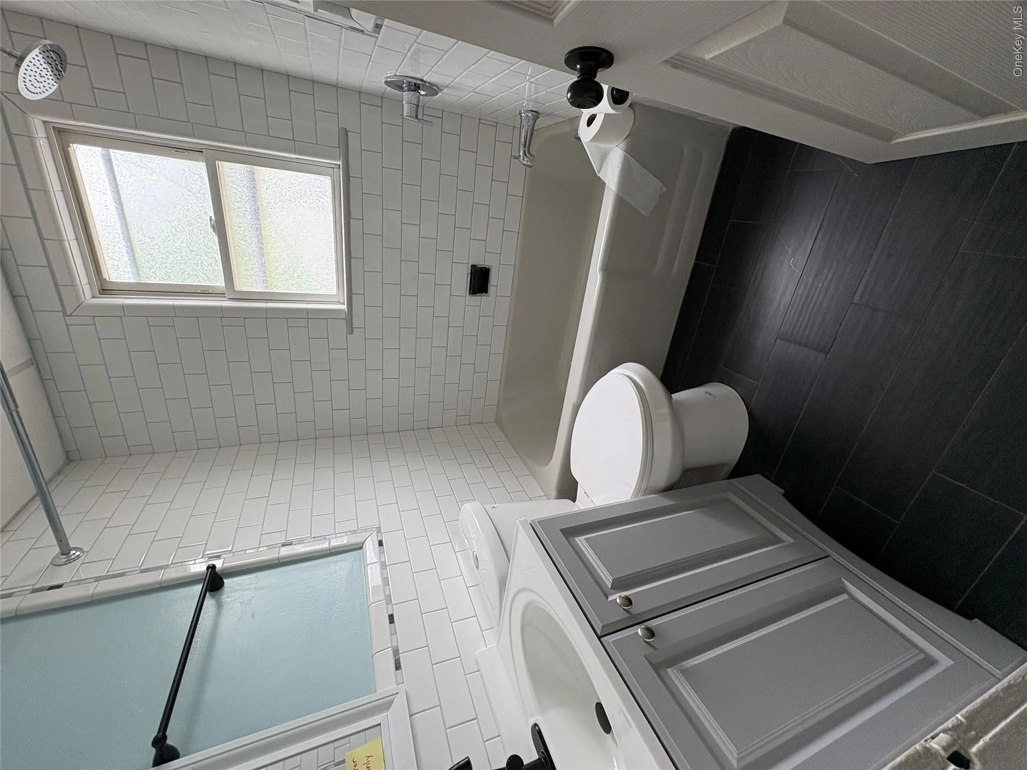 Bathroom featuring white subway tile surround,flooring, a light gray vanity with cabinet storage, a white toilet, and a white bathtub with a shower fixture Bathroom featuring white subway tile surround,flooring, a light gray vanity with cabinet storage, a white toilet, and a white bathtub with a shower fixture