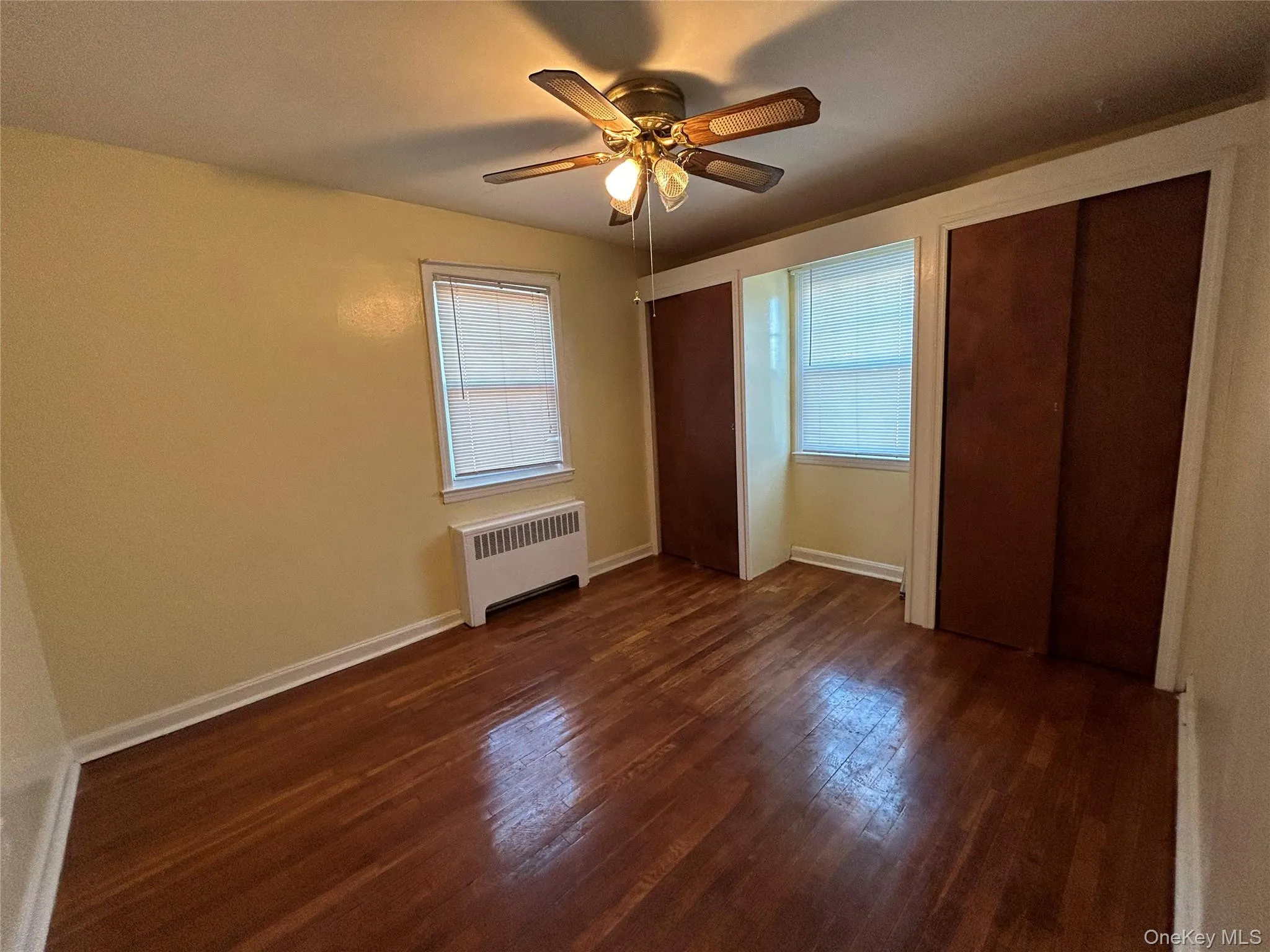 Unfurnished bedroom with baseboards, radiator, dark wood-style floors, multiple windows, and multiple closets Unfurnished bedroom with baseboards, radiator, dark wood-style floors, multiple windows, and multiple closets