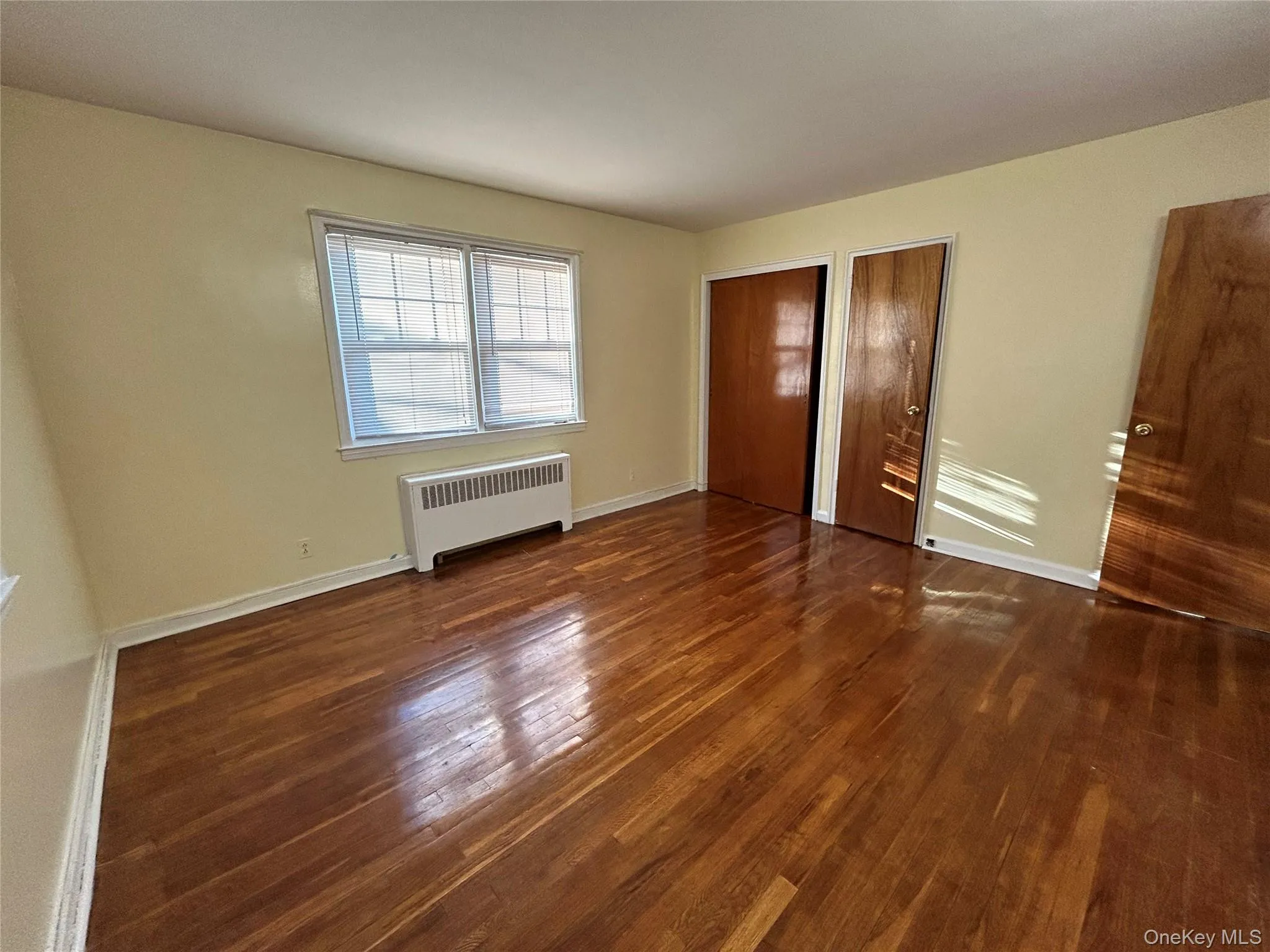 Unfurnished bedroom featuring radiator heating unit, two closets, dark wood finished floors, and baseboards Unfurnished bedroom featuring radiator heating unit, two closets, dark wood finished floors, and baseboards