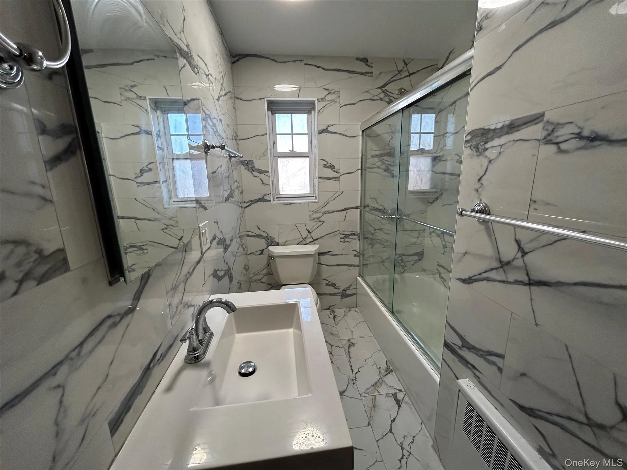 Full bathroom with marble finish floor, shower / bath combination with glass door, toilet, vanity, and stone wall Full bathroom with marble finish floor, shower / bath combination with glass door, toilet, vanity, and stone wall