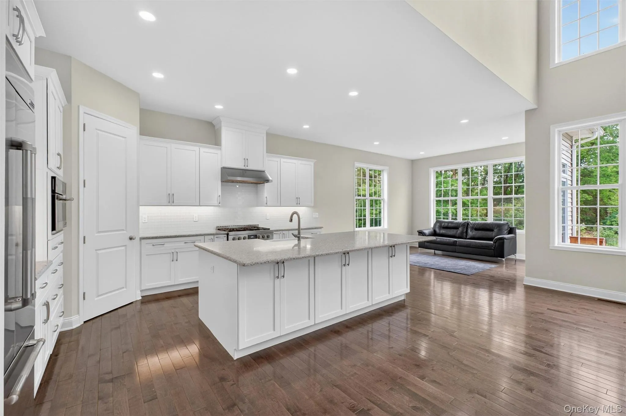 Spacious open-concept living area featuring rich wood-finish flooring, a contemporary kitchen island with quartz countertop, white shaker-style cabinetry, and abundant natural light through multiple windows Spacious open-concept living area featuring rich wood-finish flooring, a contemporary kitchen island with quartz countertop, white shaker-style cabinetry, and abundant natural light through multiple windows