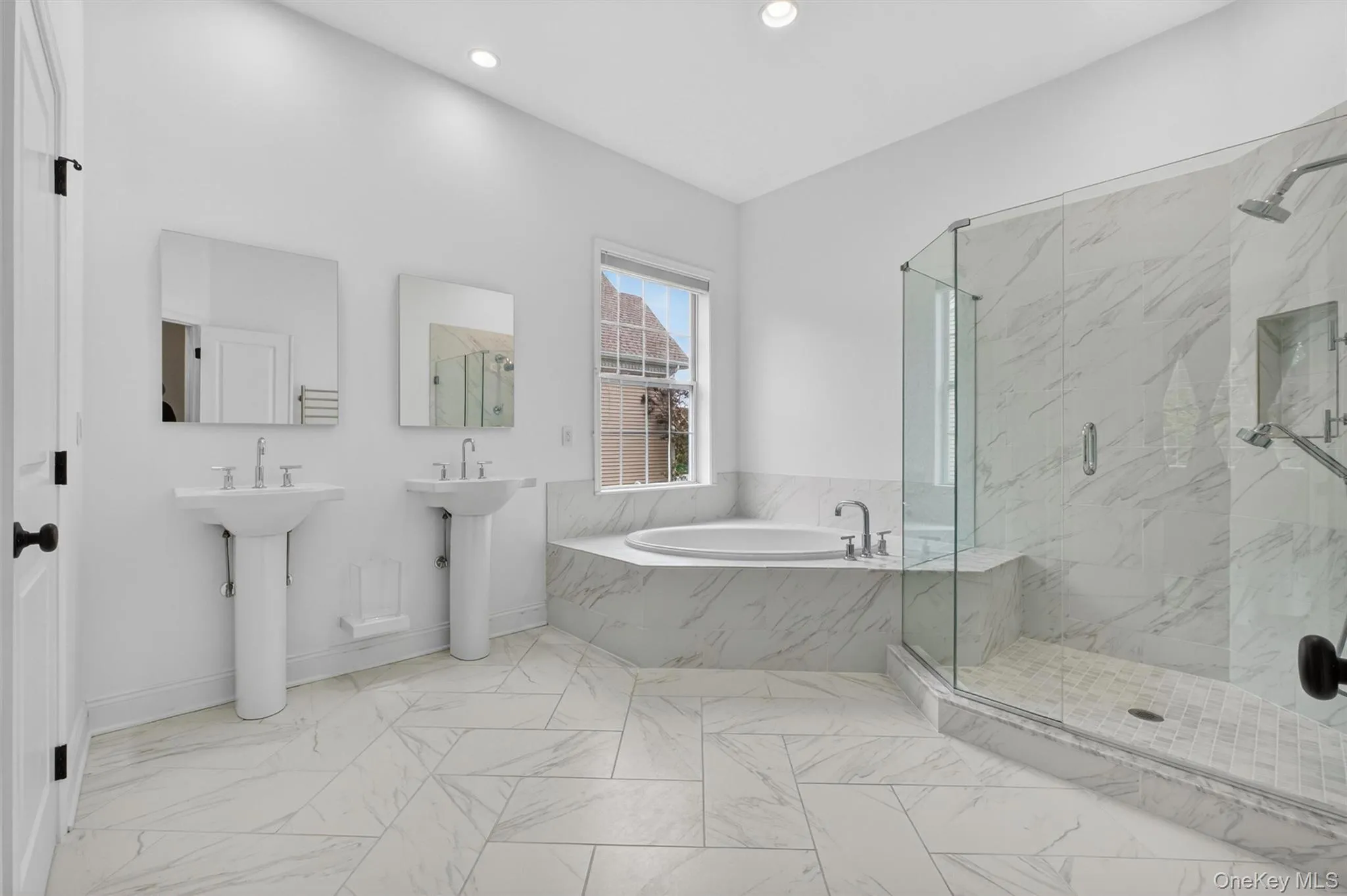 Spacious primary bathroom featuring marble-look tile flooring, dual pedestal sinks with mirrors, a soaking tub with surrounding marble-look accents, and a glass-enclosed shower with a built-in bench Spacious primary bathroom featuring marble-look tile flooring, dual pedestal sinks with mirrors, a soaking tub with surrounding marble-look accents, and a glass-enclosed shower with a built-in bench