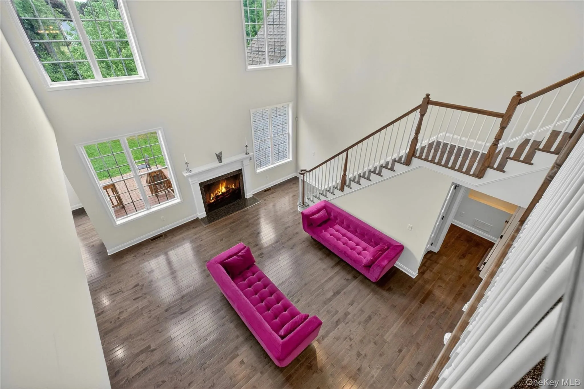 Two-story living area featuring hardwood flooring, a fireplace with a white mantel, multiple large windows, and a hardwood staircase with white balusters Two-story living area featuring hardwood flooring, a fireplace with a white mantel, multiple large windows, and a hardwood staircase with white balusters