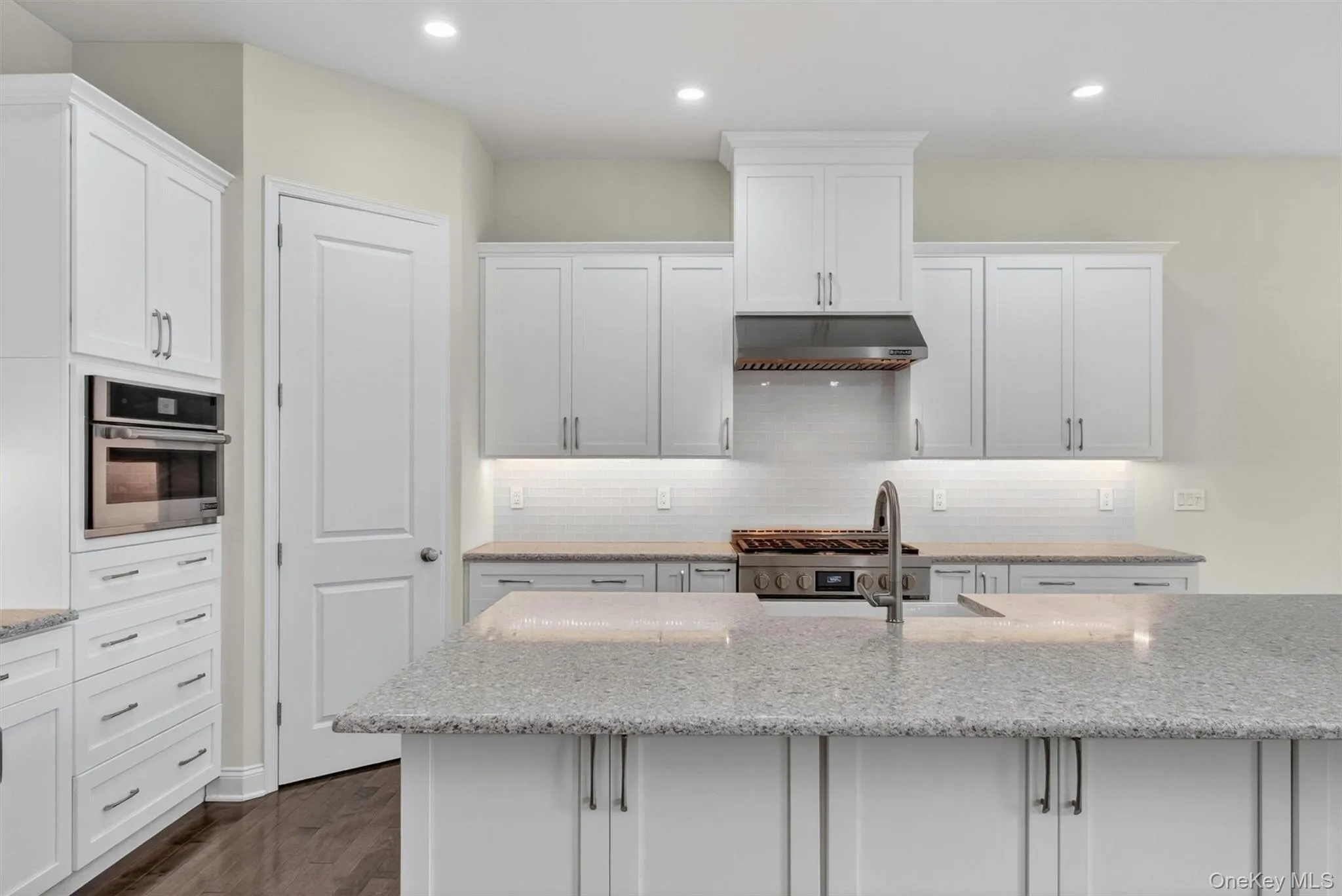 Contemporary kitchen featuring white shaker cabinetry, stainless steel appliances including a built-in oven and range hood, light-toned speckled countertops, and a subway tile backsplash Contemporary kitchen featuring white shaker cabinetry, stainless steel appliances including a built-in oven and range hood, light-toned speckled countertops, and a subway tile backsplash