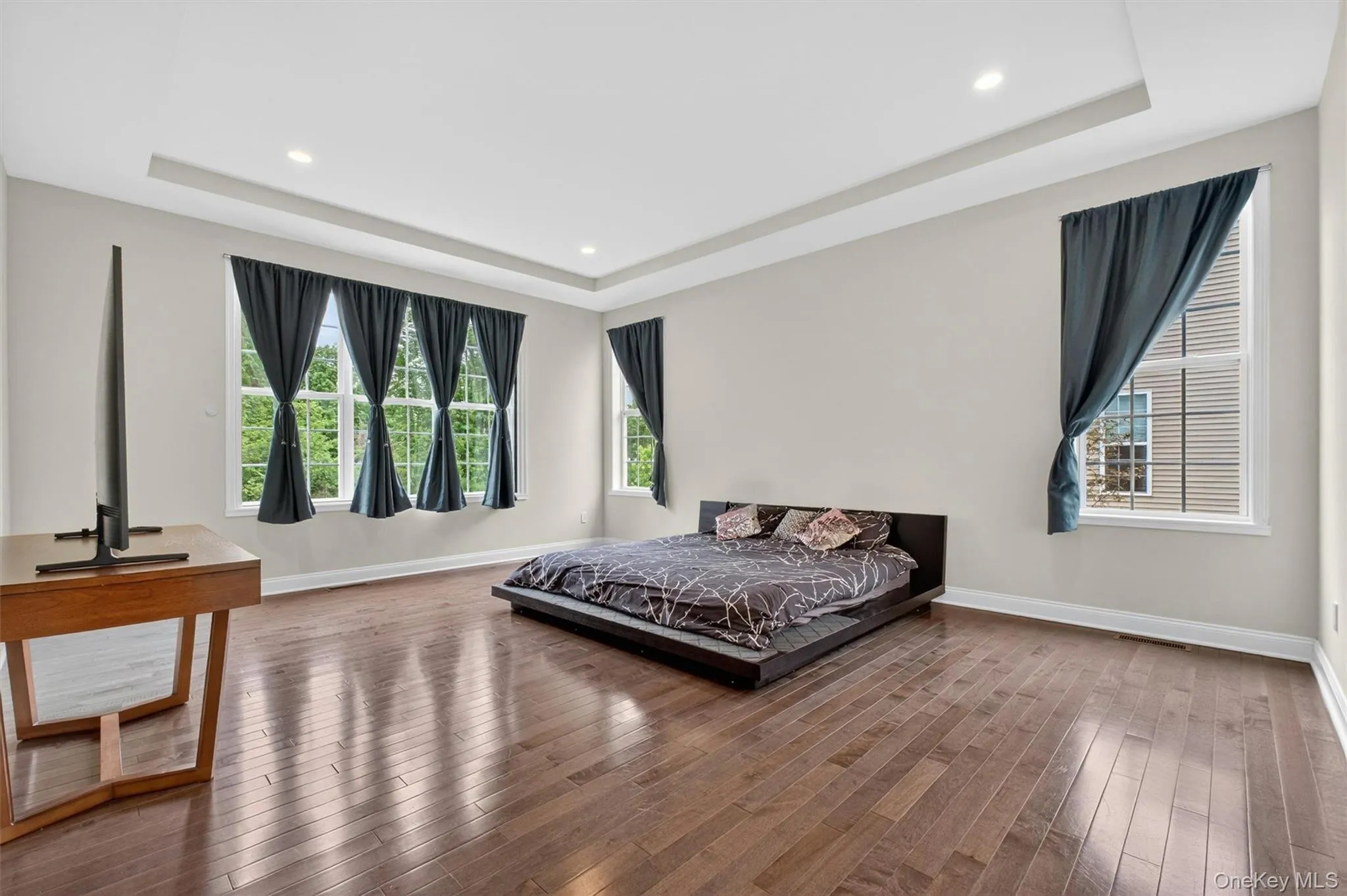 Primary bedroom featuring rich wood-finish flooring, a tray ceiling with recessed lighting, and multiple windows with white trim Primary bedroom featuring rich wood-finish flooring, a tray ceiling with recessed lighting, and multiple windows with white trim