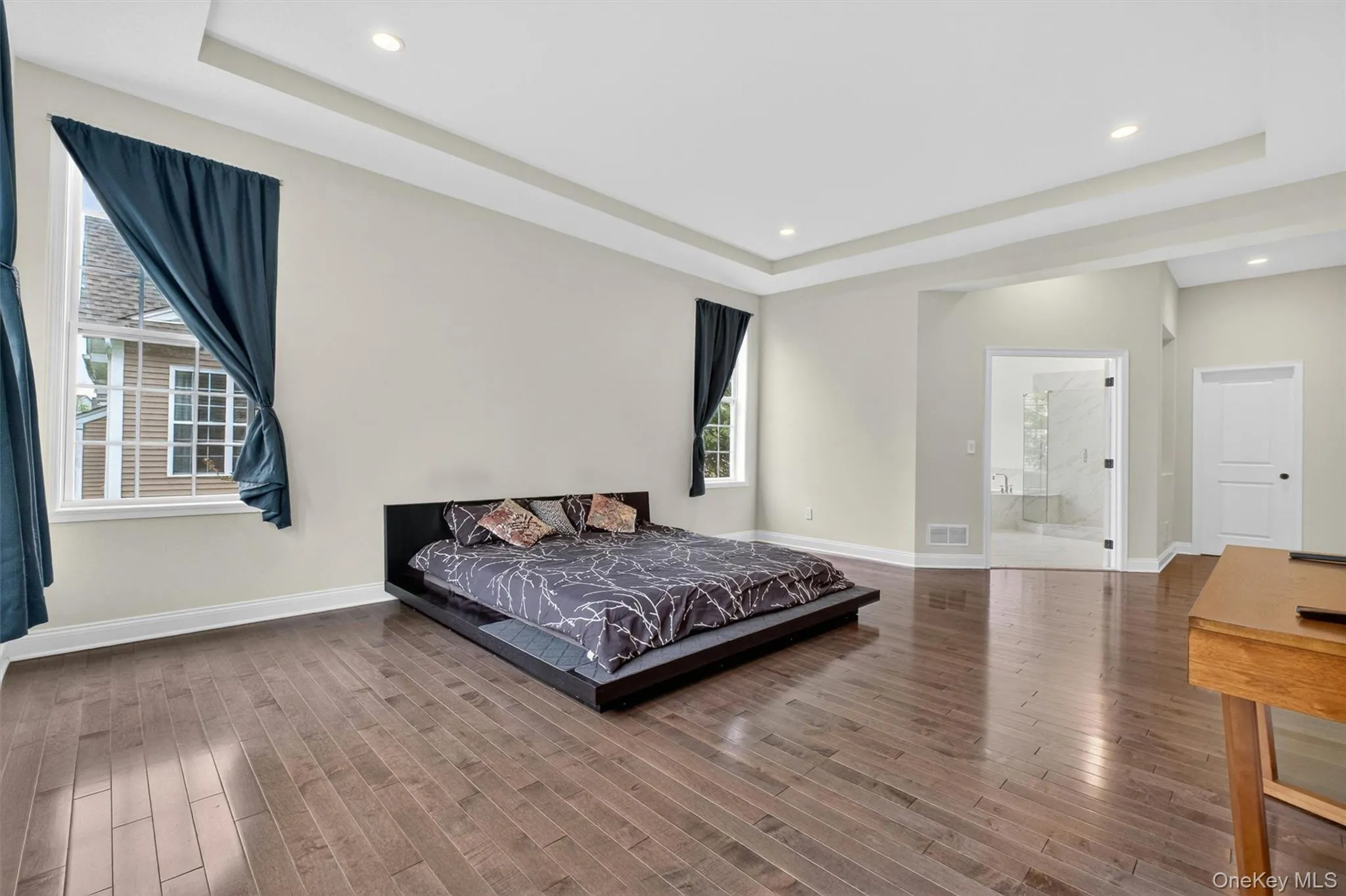 Spacious primary en suite featuring dark wood-finish flooring, recessed lighting, tray ceiling, and an en-suite bathroom with a glass-enclosed shower Spacious primary en suite featuring dark wood-finish flooring, recessed lighting, tray ceiling, and an en-suite bathroom with a glass-enclosed shower