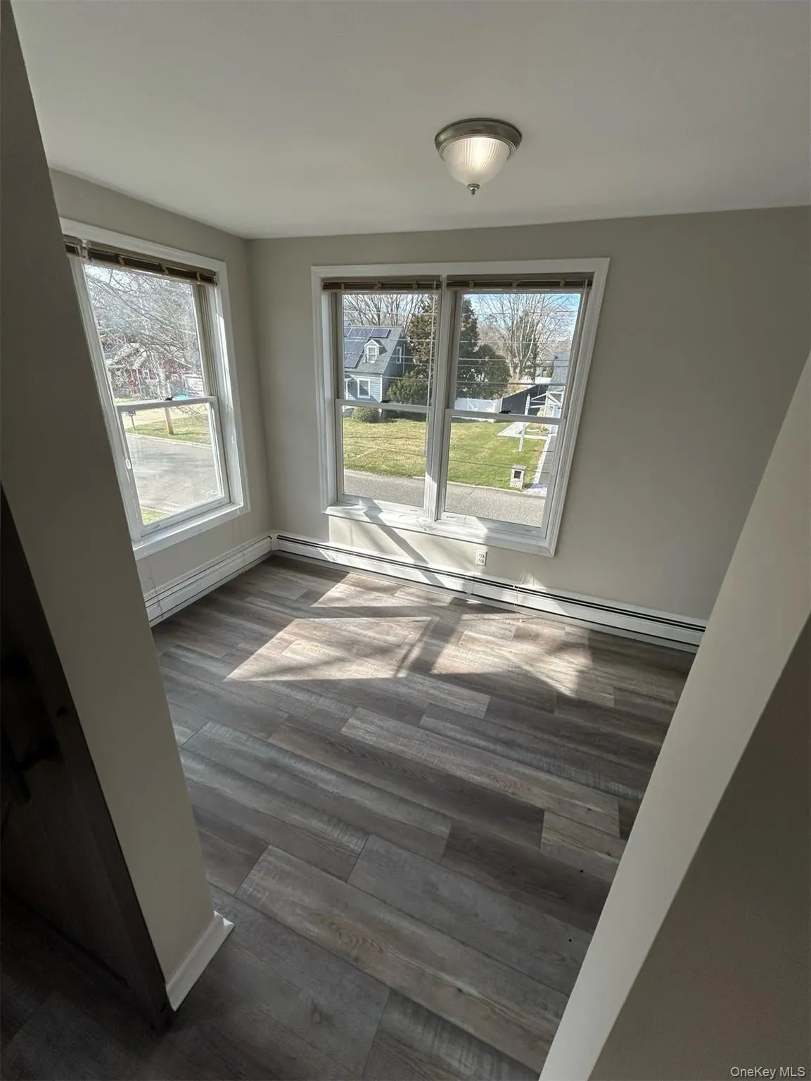 Room featuring three windows with white trim, wood-finish flooring, light gray wall paint, and a ceiling-mounted light fixture Room featuring three windows with white trim, wood-finish flooring, light gray wall paint, and a ceiling-mounted light fixture