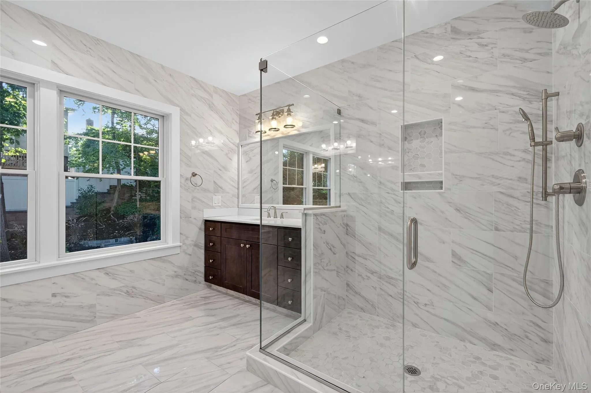 Bathroom featuring healthy amount of natural light, a marble finish shower, light marble finish floors, vanity, and tile walls Bathroom featuring healthy amount of natural light, a marble finish shower, light marble finish floors, vanity, and tile walls