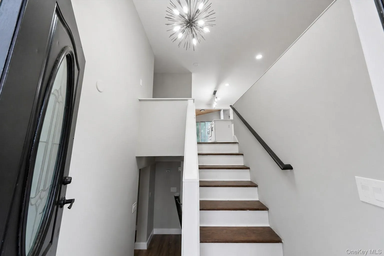 Foyer entry with a decorative glass-paneled door, contemporary sputnik chandelier, recessed lighting, and a staircase featuring dark wood-finish treads and a black handrail Foyer entry with a decorative glass-paneled door, contemporary sputnik chandelier, recessed lighting, and a staircase featuring dark wood-finish treads and a black handrail